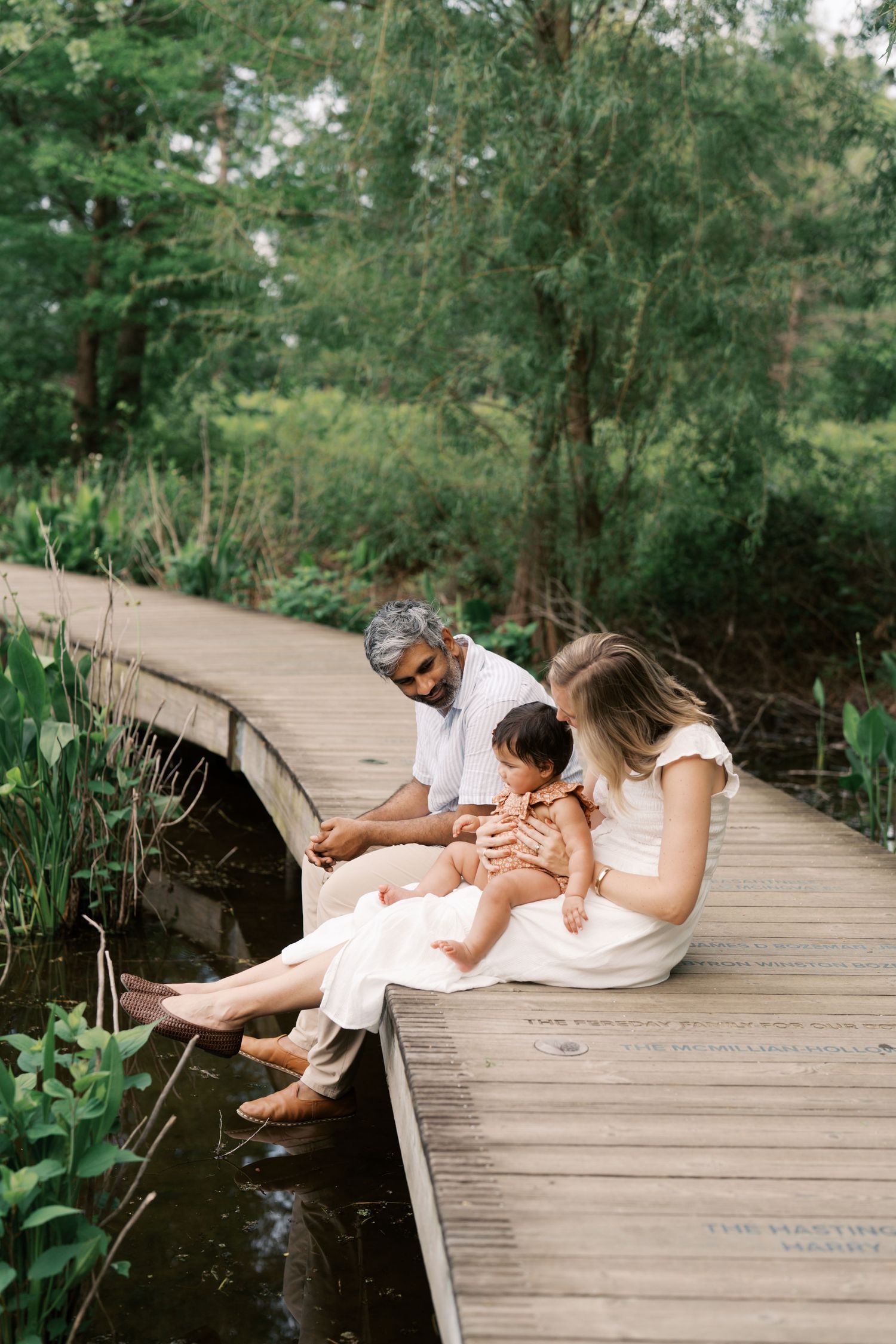 Family with baby sitting on wooden bridge over pond surrounded by lush greenery.