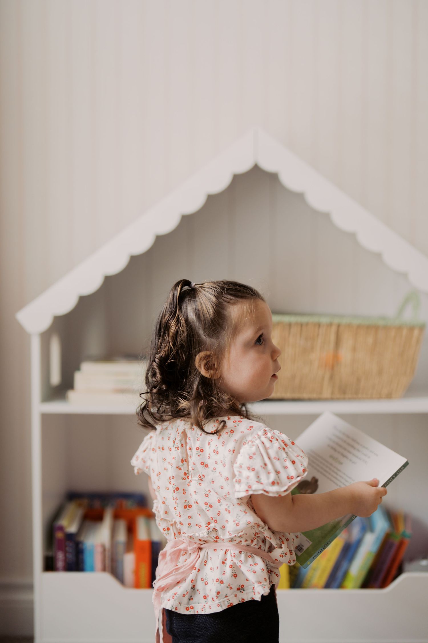 Small child in floral print top reading a book in front of a white bookshelf with scalloped trim detail.