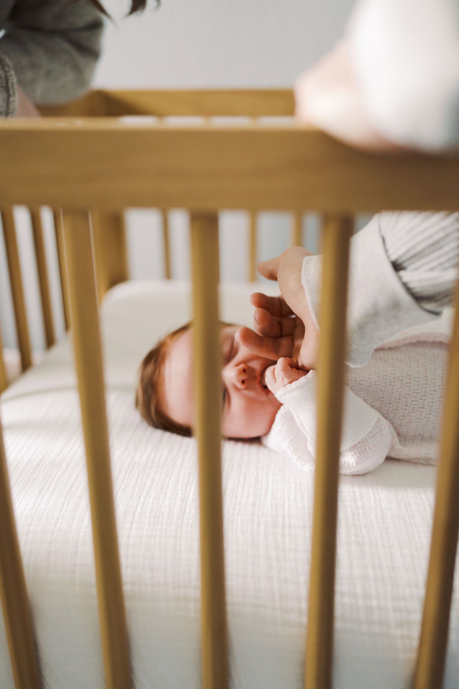 A newborn baby lying in a wooden crib, reaching up with tiny hands toward a parent standing nearby.