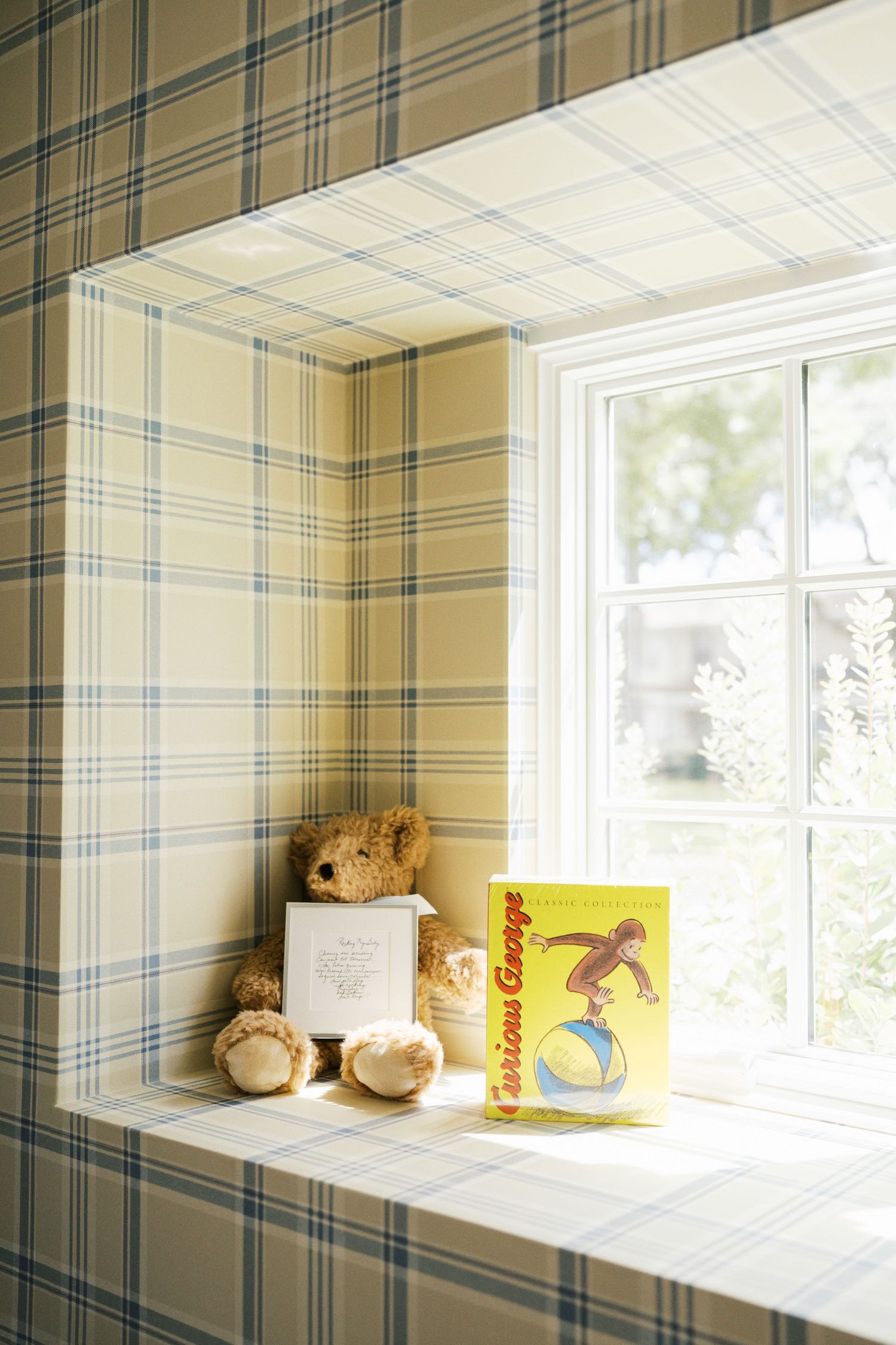 Cozy window seat nook with plaid wallpaper, teddy bear, and Curious George book in a charming children's room.