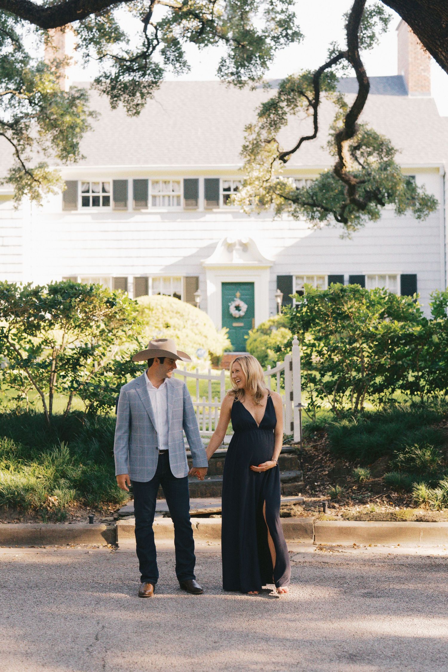 Pregnant couple holding hands in front of white house, man in cowboy hat and blazer, woman in black maxi dress.
