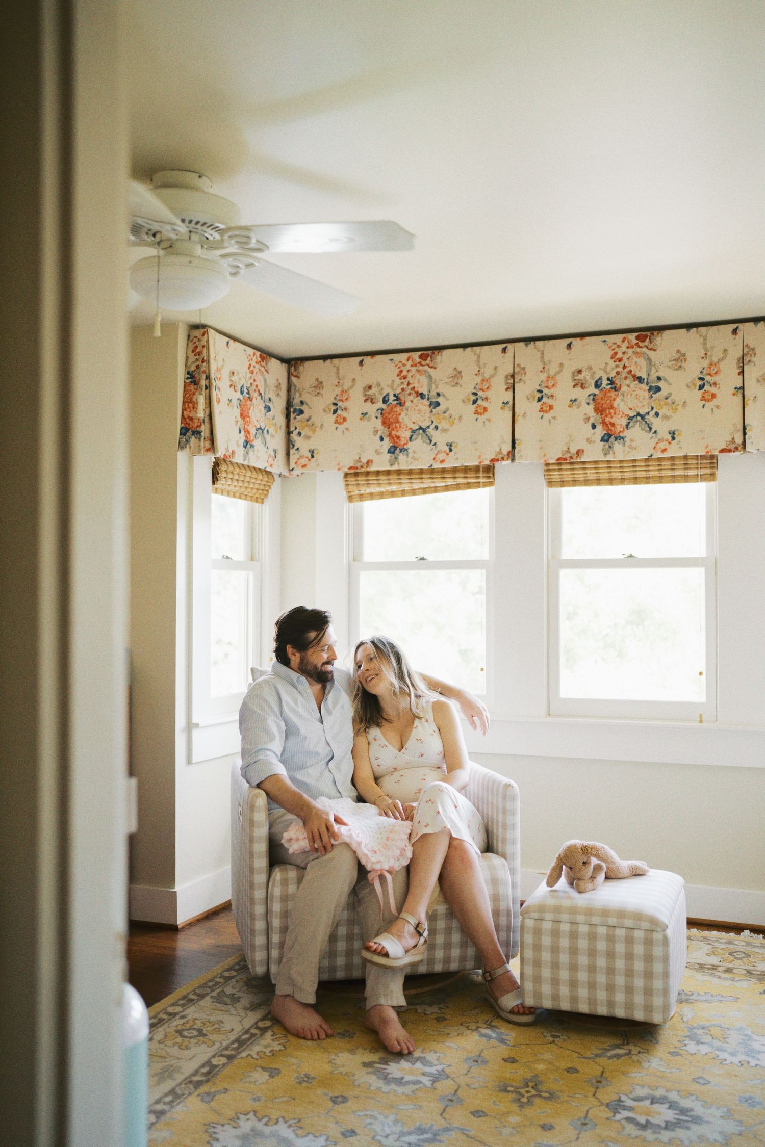 Couple relaxing together in cozy nursery chair with floral valances and small dog nearby.