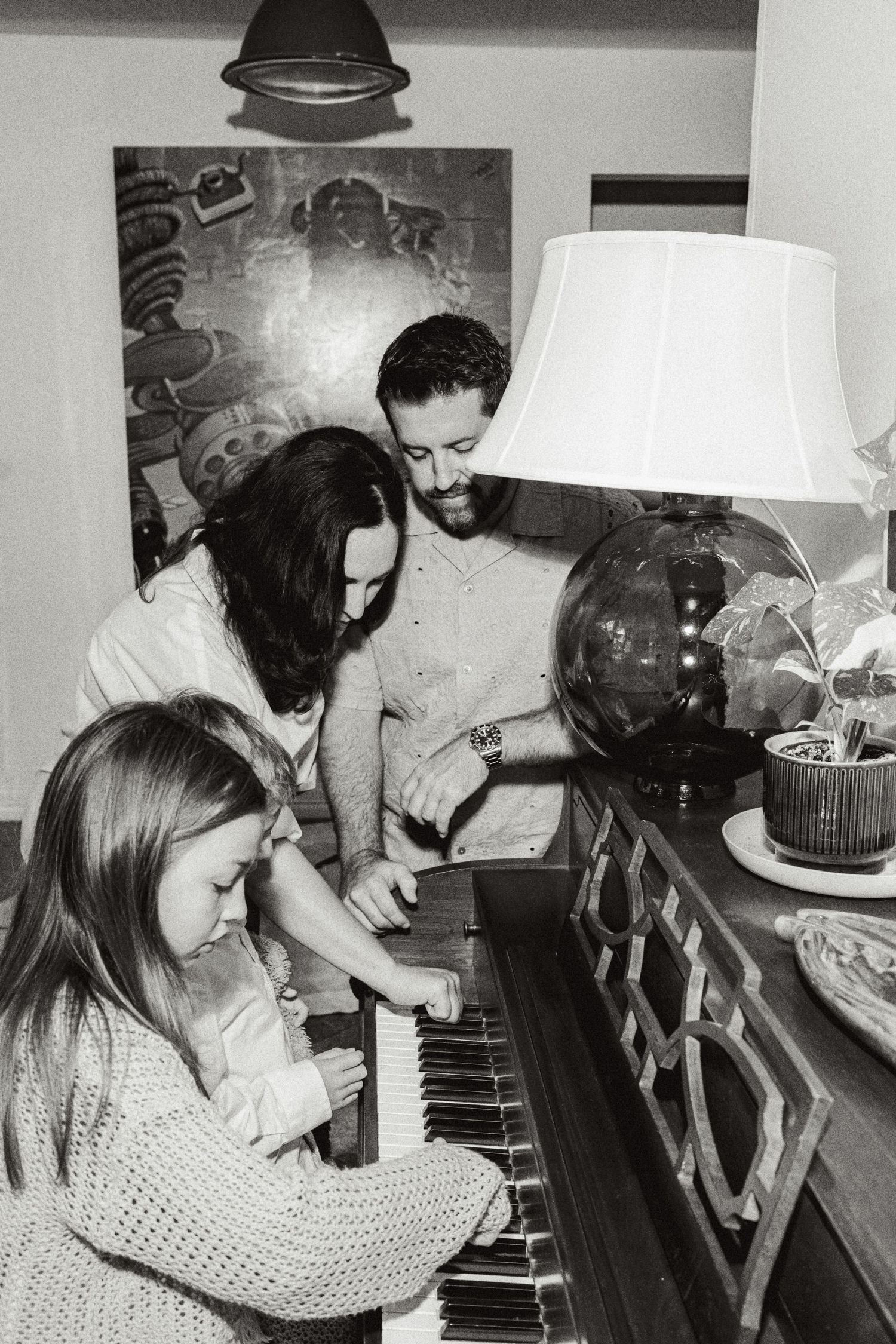 Black and white photo of a family gathered around a piano, with a young girl playing while two adults look on.