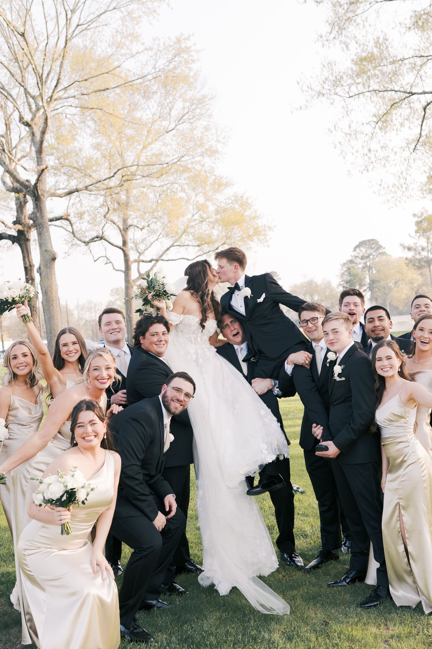 Joyful wedding party posing together on a lawn with bridesmaids in champagne dresses and groomsmen in black tuxedos.