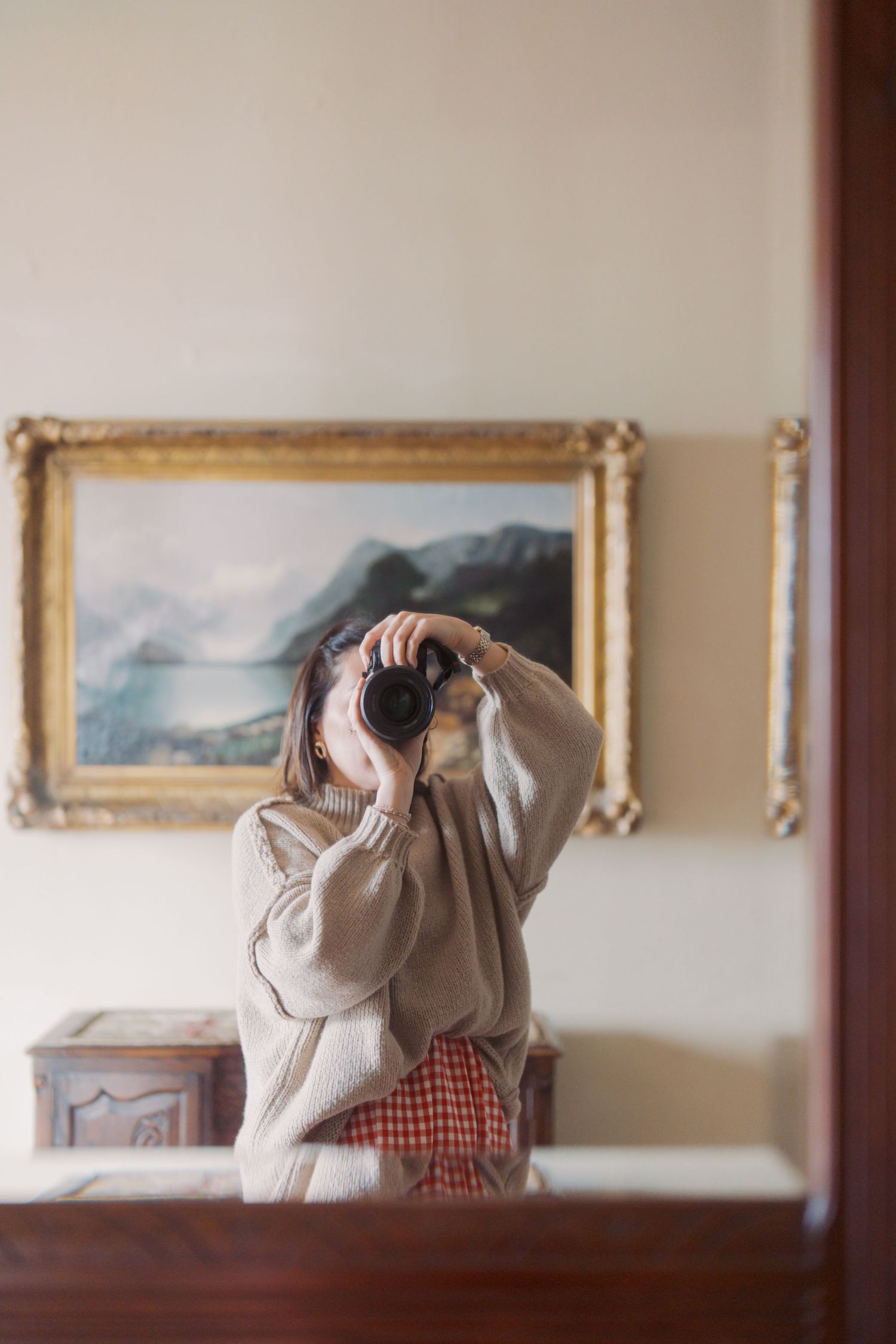 Woman taking a mirror selfie with a DSLR camera, wearing a beige sweater, with a framed landscape painting on the wall behind her.