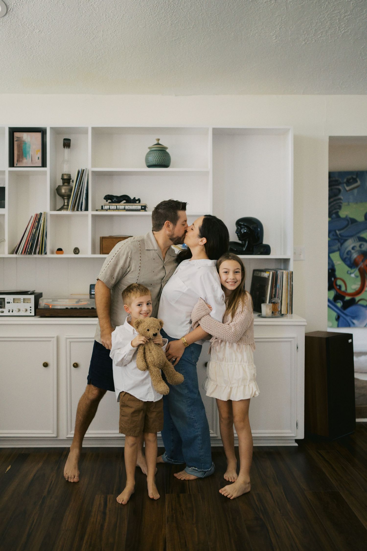 A happy family of four poses indoors, parents kissing while children hold a teddy bear in a cozy living room.