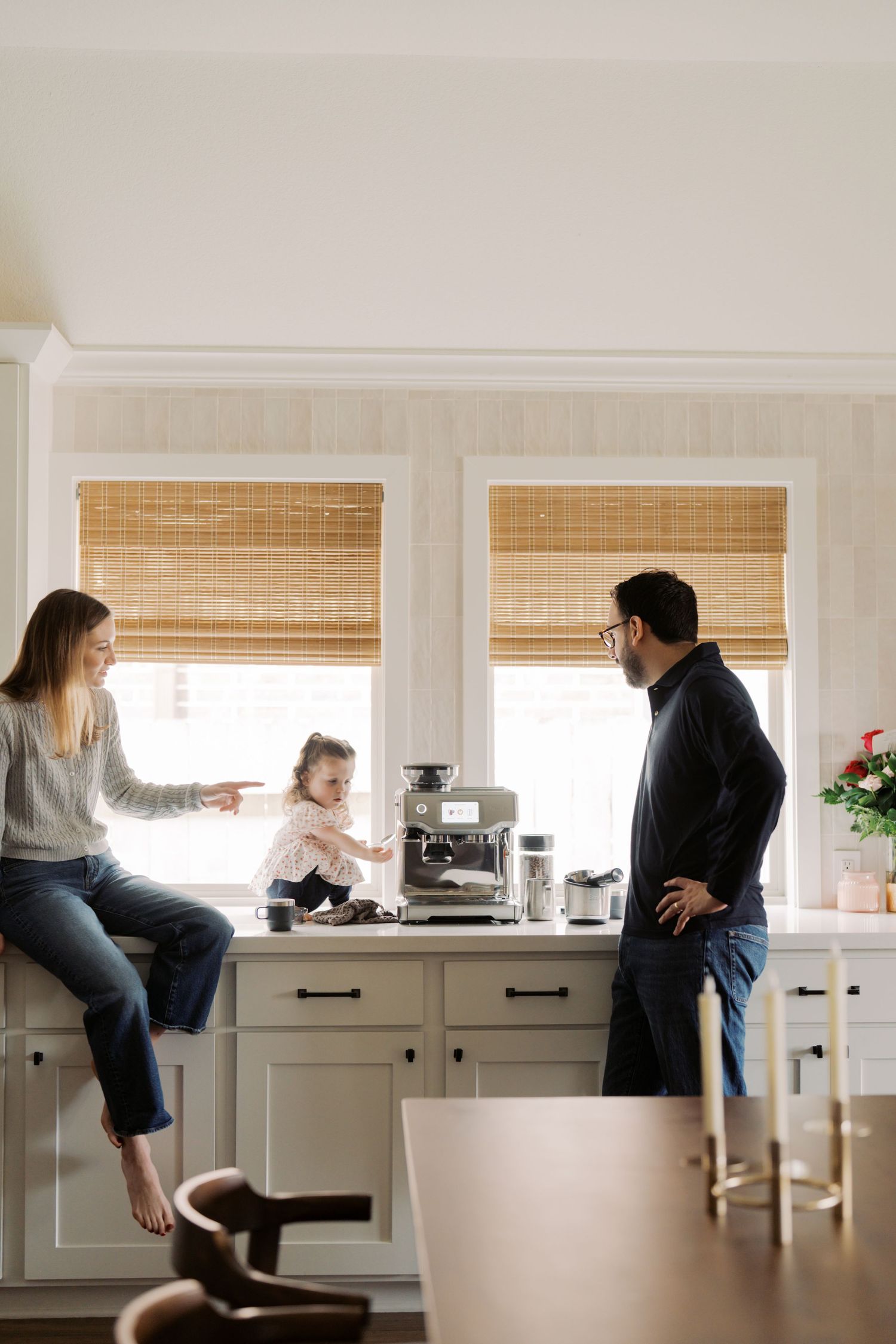 A family of three - a mother, father, and young child - interacting in a cozy, well-lit kitchen.