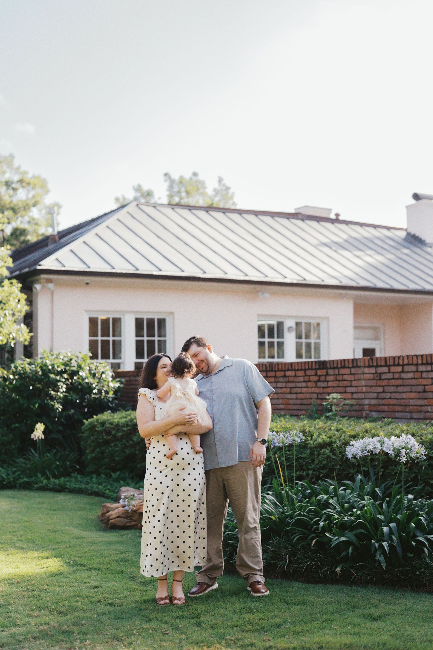 A couple stands on green lawn holding a newborn baby in front of a charming white house with brick accents.