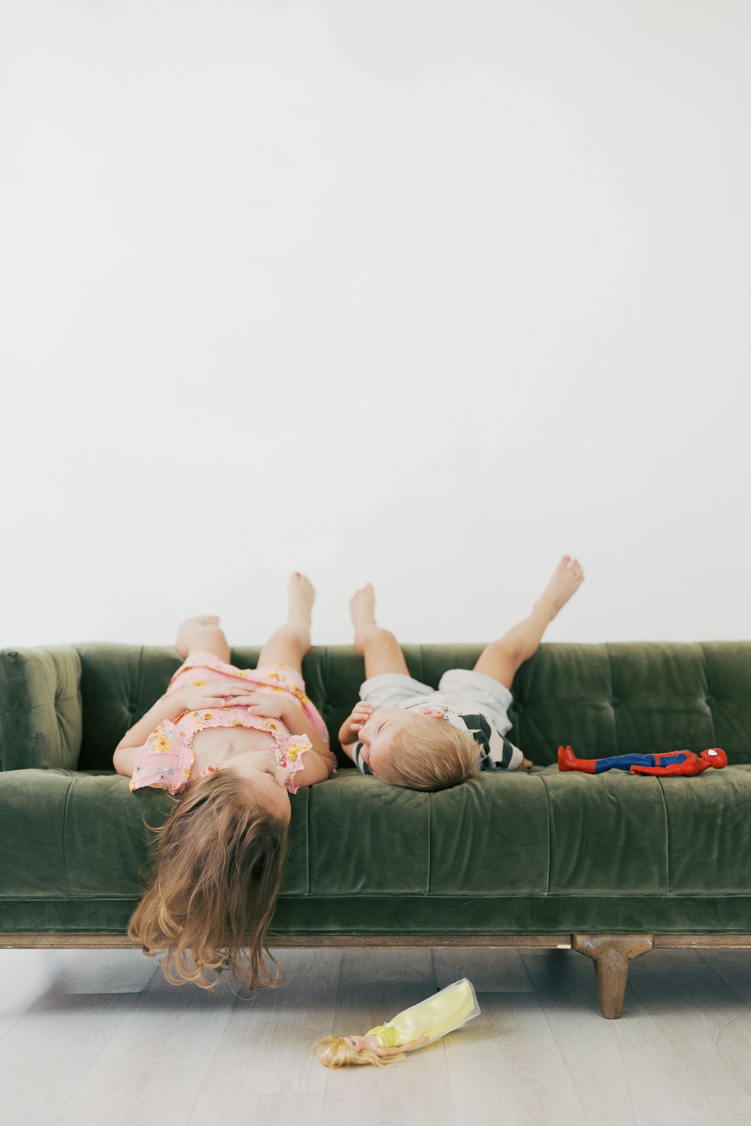 Two children laying upside down on a plush green velvet sofa with toys scattered nearby.