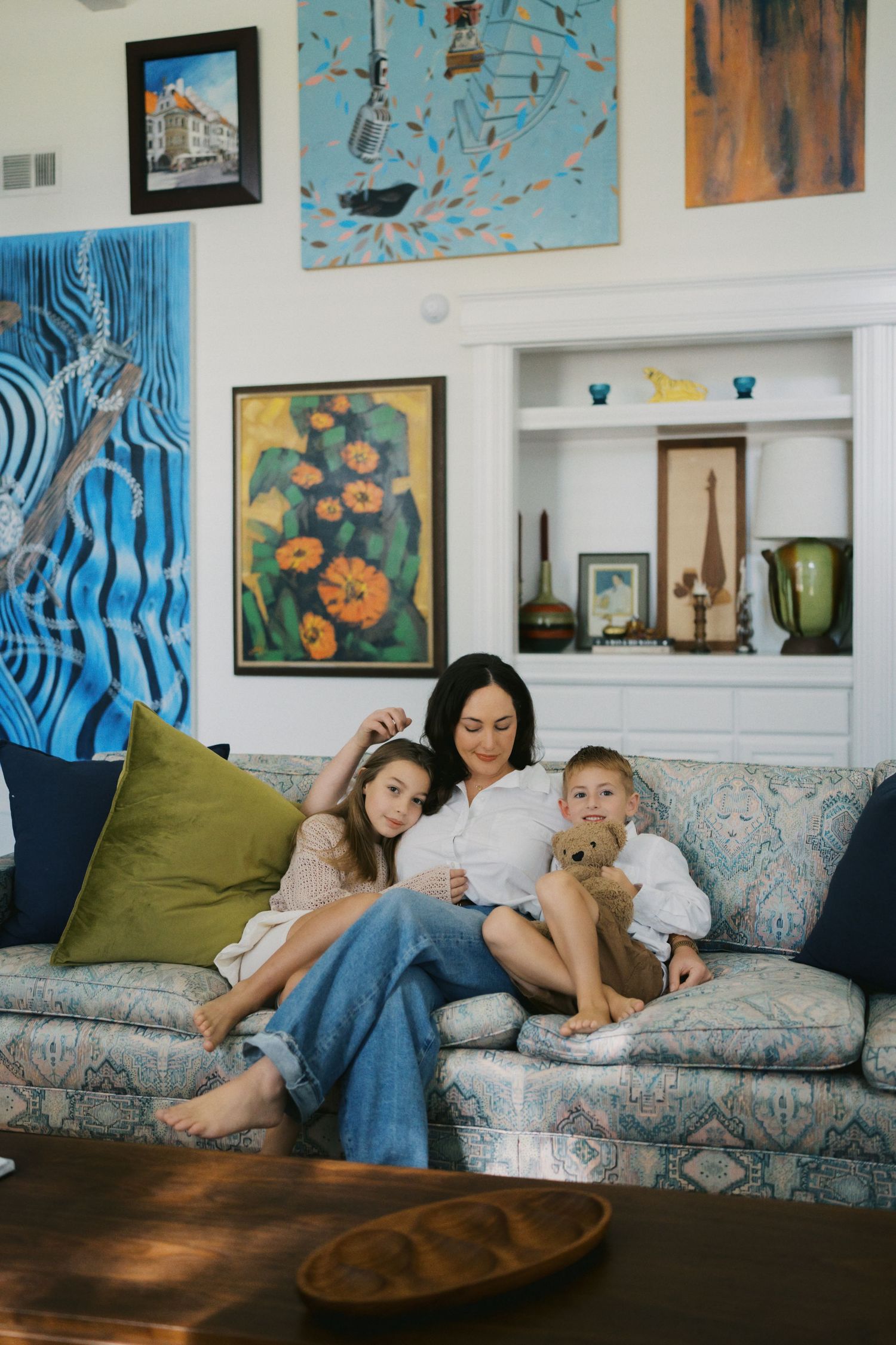 A mother and two children relax together on a floral sofa surrounded by colorful artwork in a bright, eclectic living room.