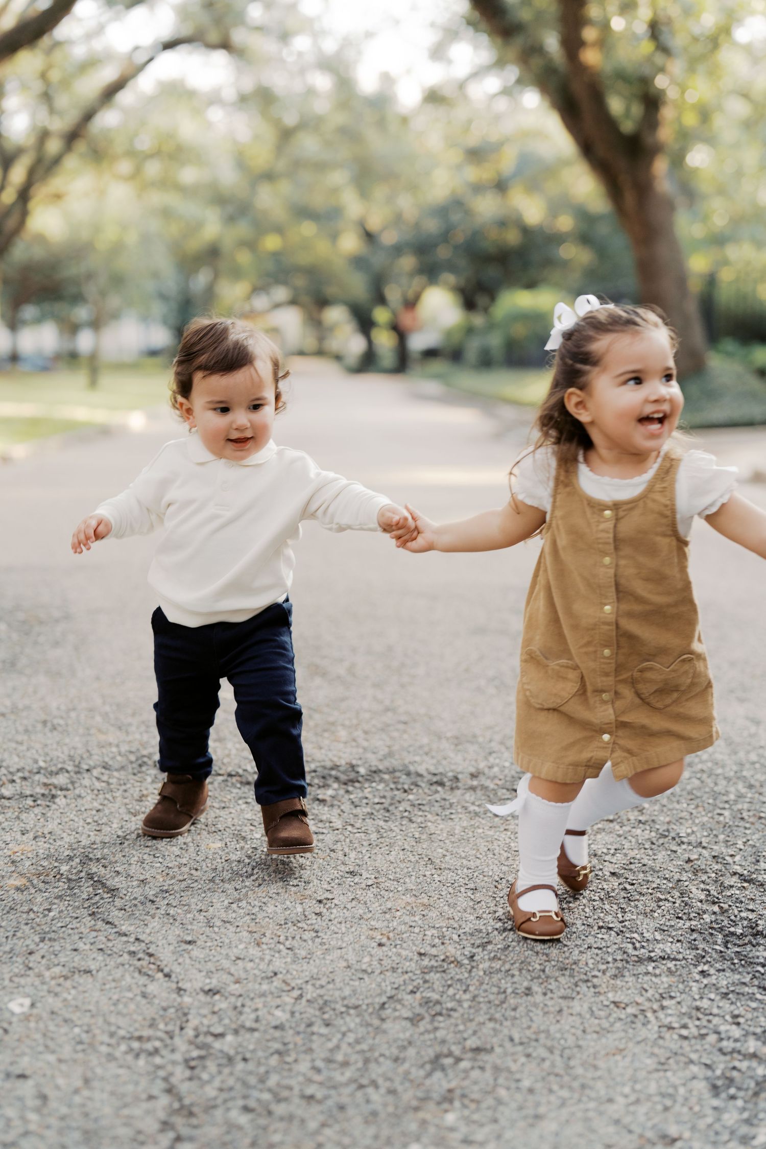 Two young children holding hands and running together on a path surrounded by trees in a park.