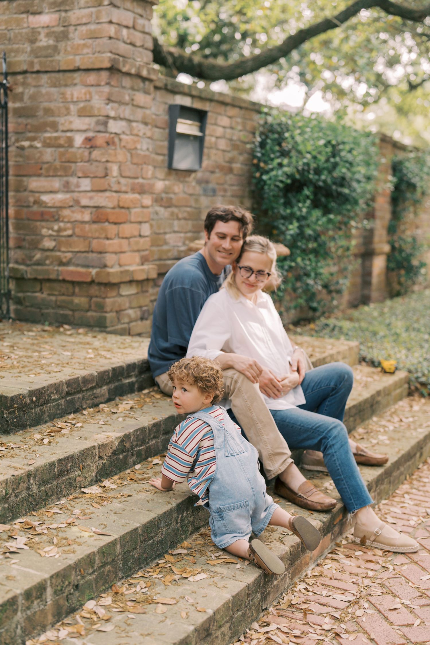 Family of three sitting on stone steps outdoors, parents smiling with toddler in denim overalls by brick wall.