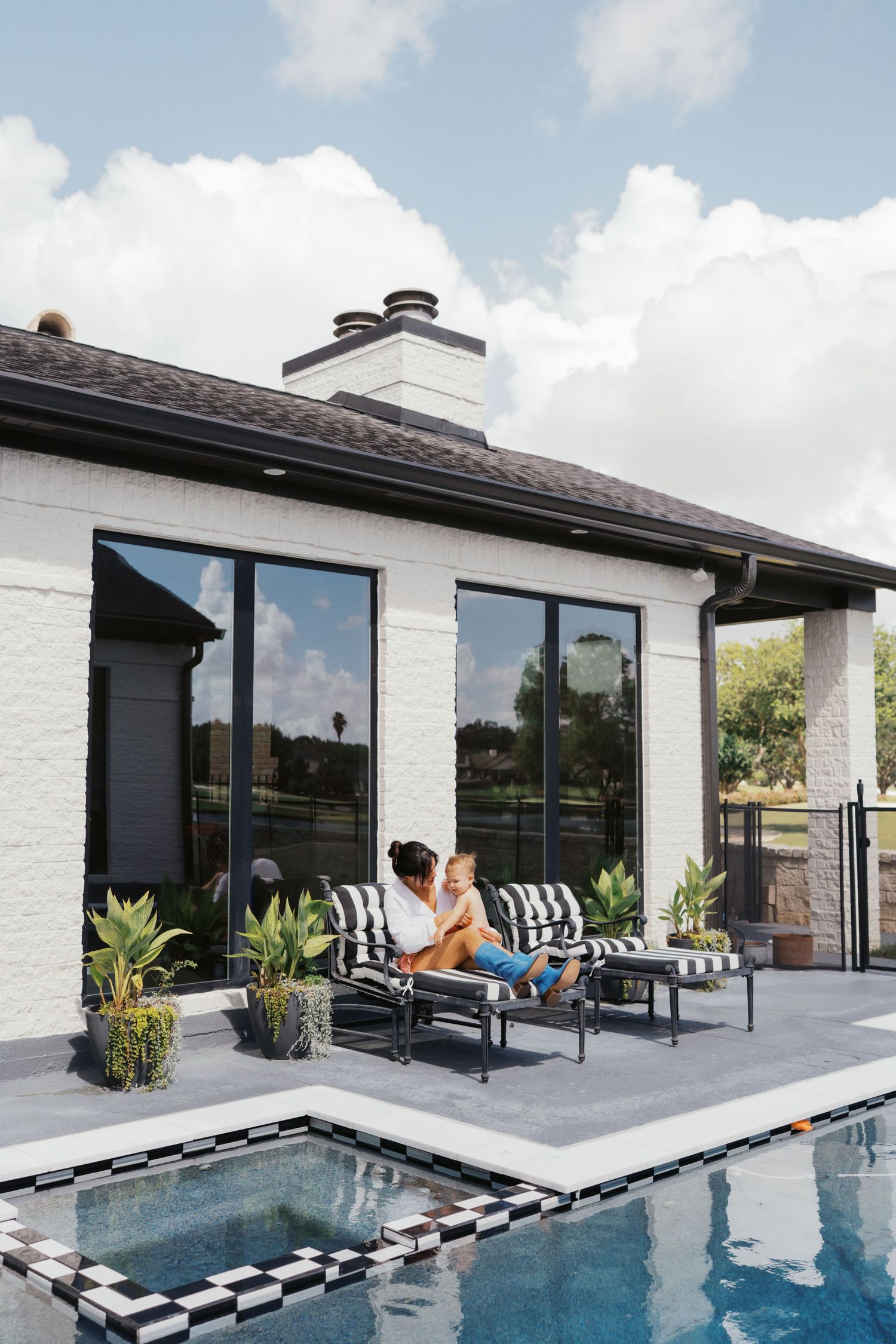 A woman and child relax on striped patio furniture beside a pool at a modern white brick home.