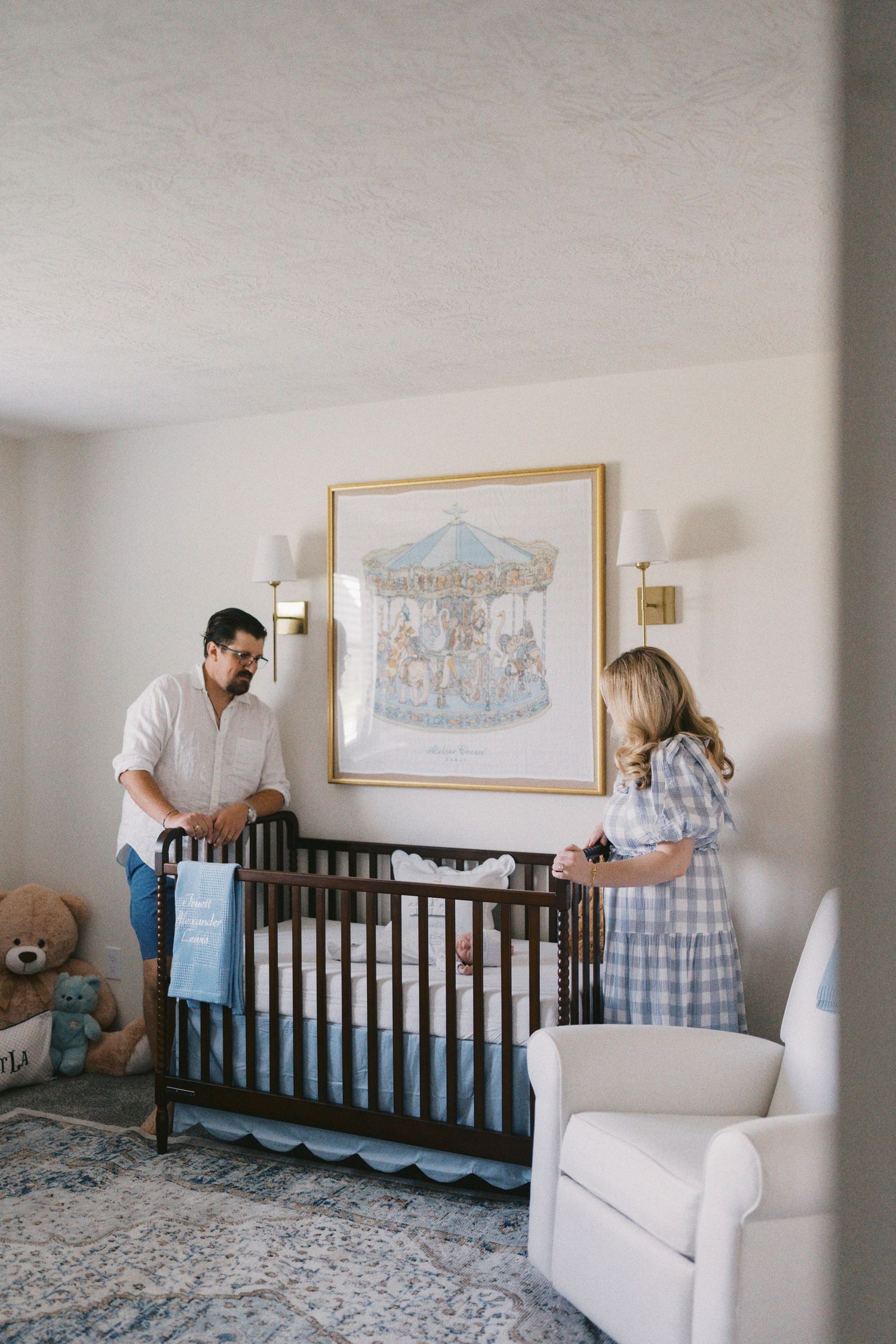 Parents arranging a dark wood crib in a blue nursery with carousel art, sconce lighting, and a white glider chair.