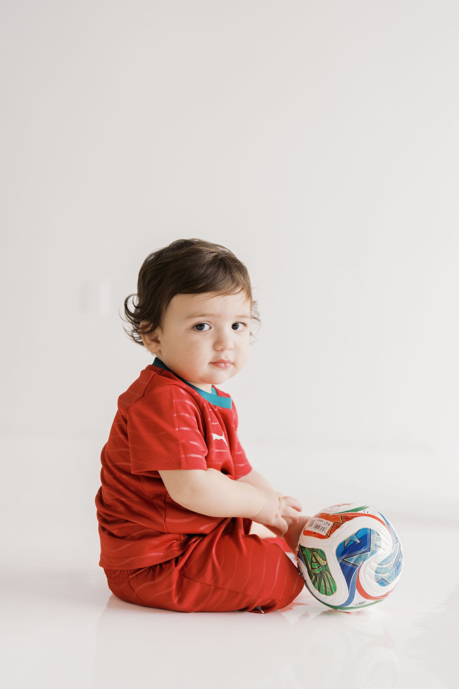 A young child in a red shirt sits on the floor and plays with a colorful ball.