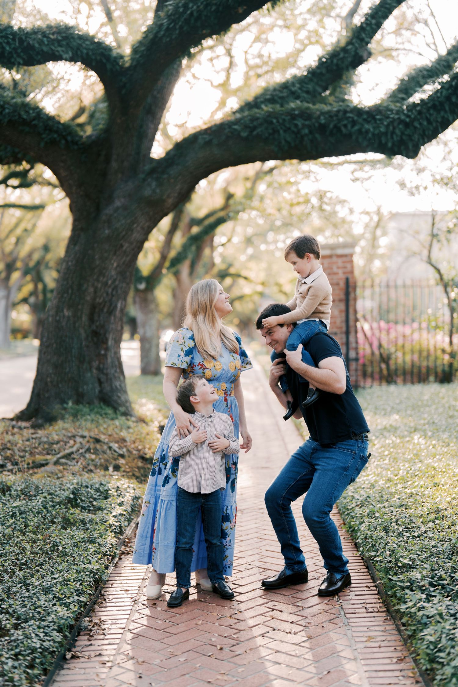 Family enjoying playful moment under large oak tree on brick pathway.