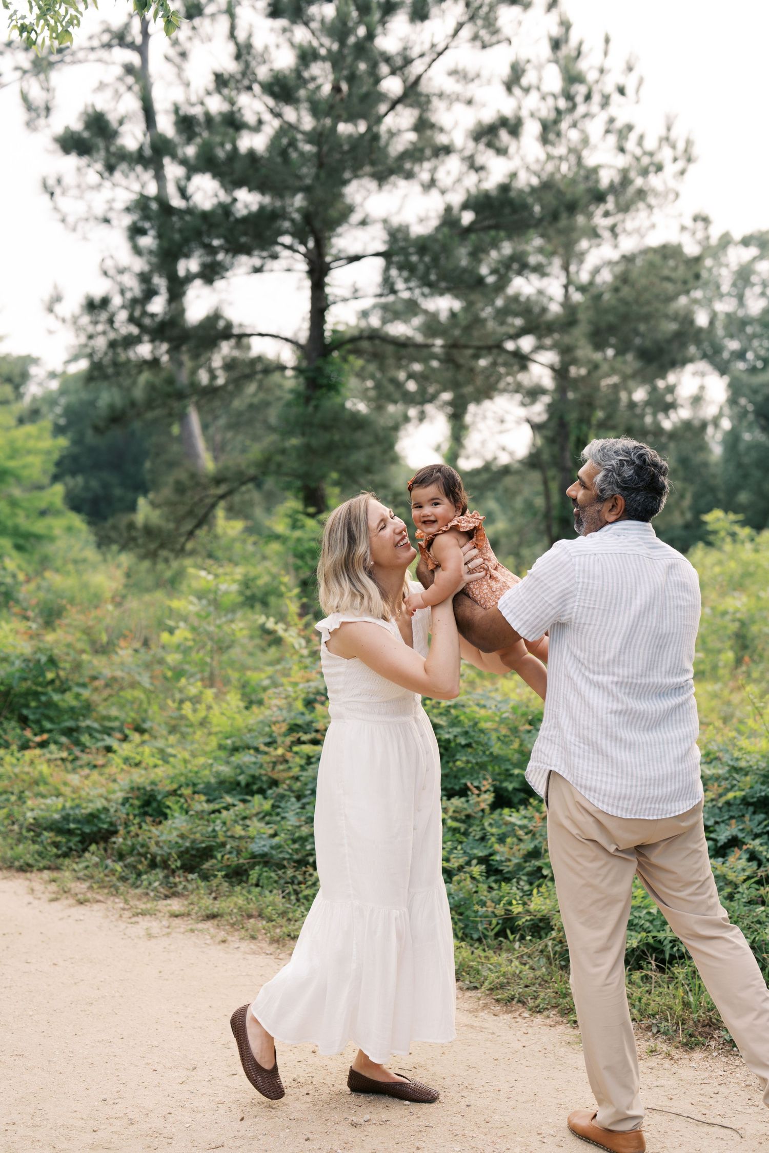 A joyful family of three plays outdoors on a wooded path, with parents lifting their laughing baby in the air.