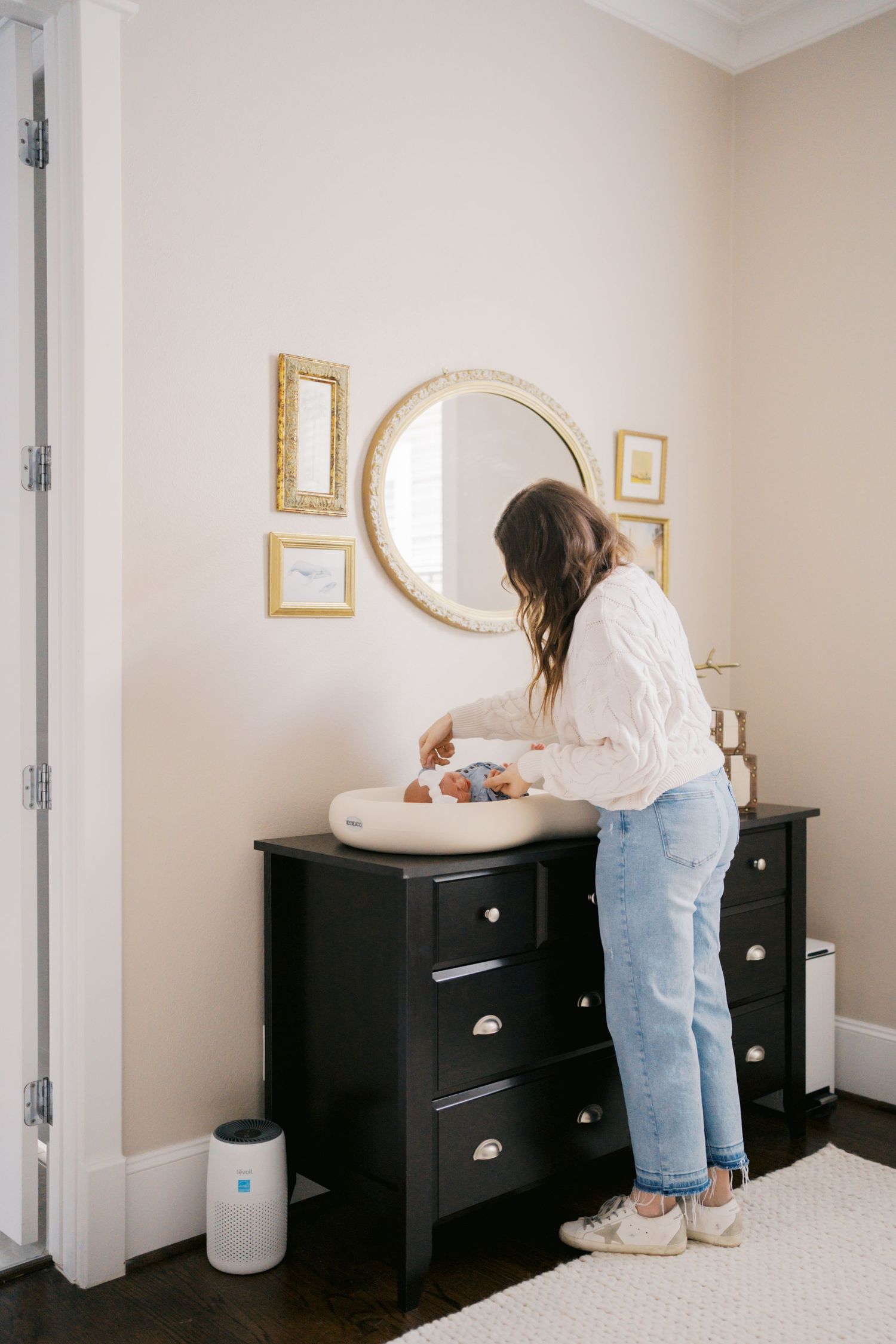 Mother changing baby on black dresser in nursery with gold mirror and frames on beige wall.