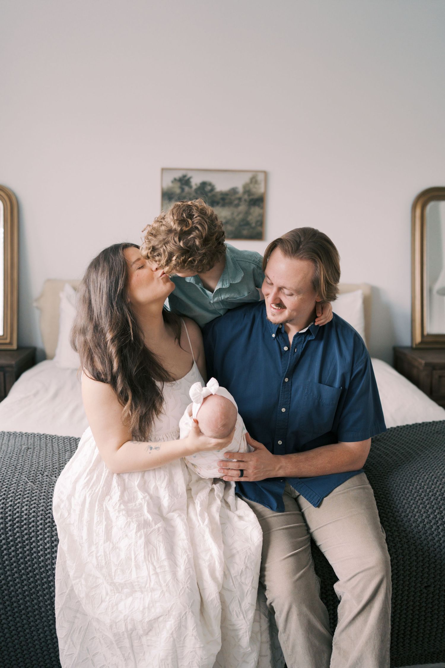 A couple sitting on a bed, smiling and kissing their newborn baby wrapped in white.