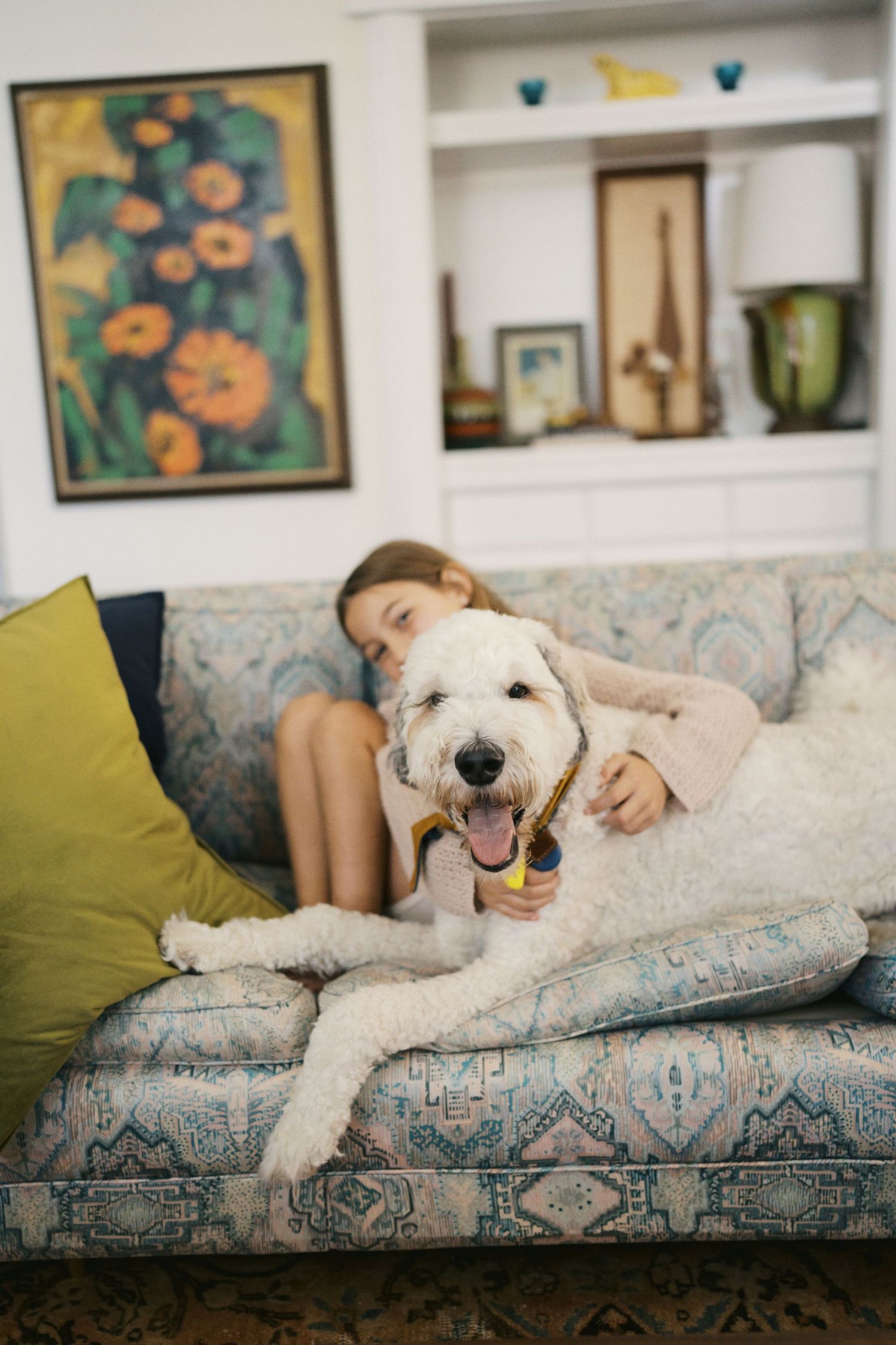Young girl relaxing on patterned sofa with large white fluffy dog holding a toy.