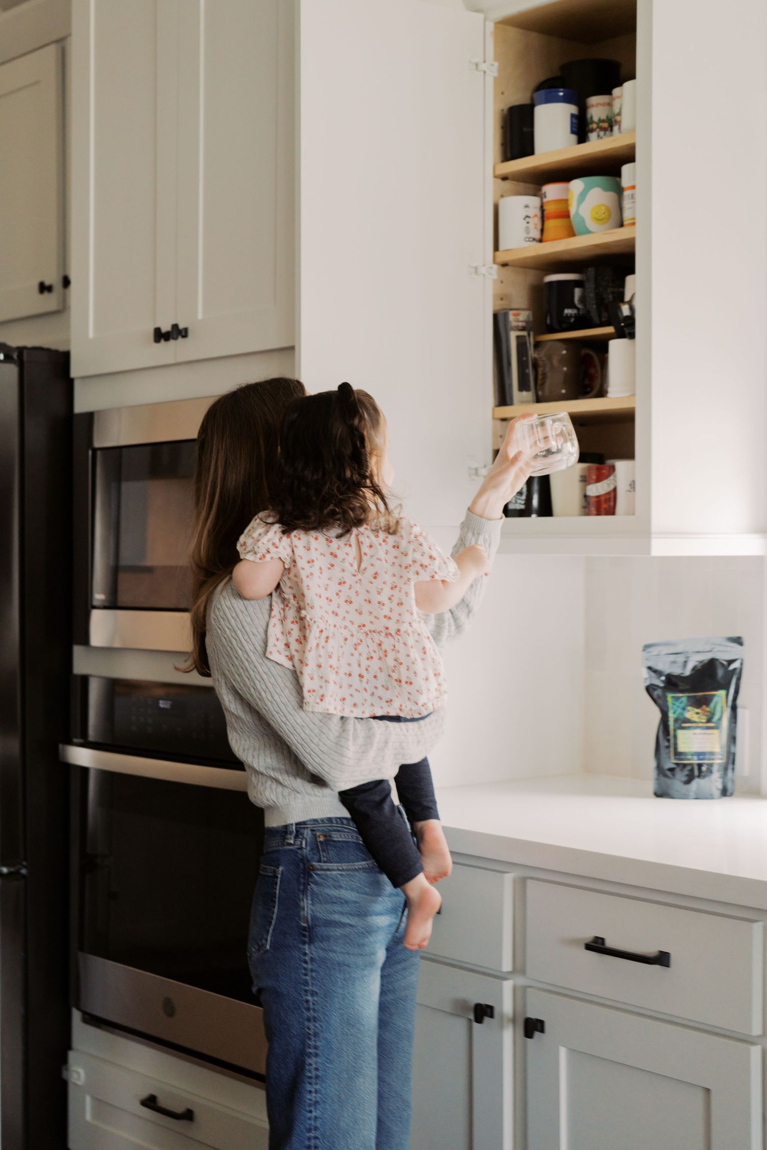 A young woman stands in a kitchen, reaching up to an open cabinet while wearing a floral blouse and jeans.