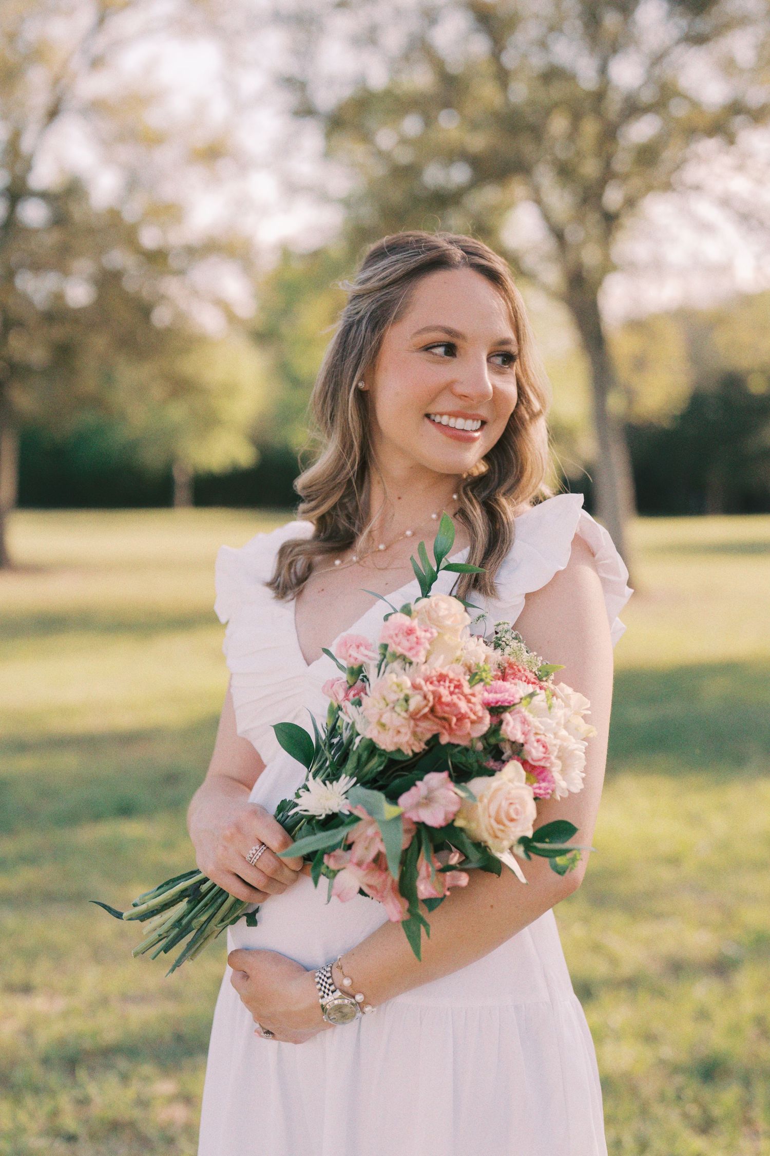 A bride in a white dress holds a pink and blush rose bouquet while standing in a sunlit field at golden hour.