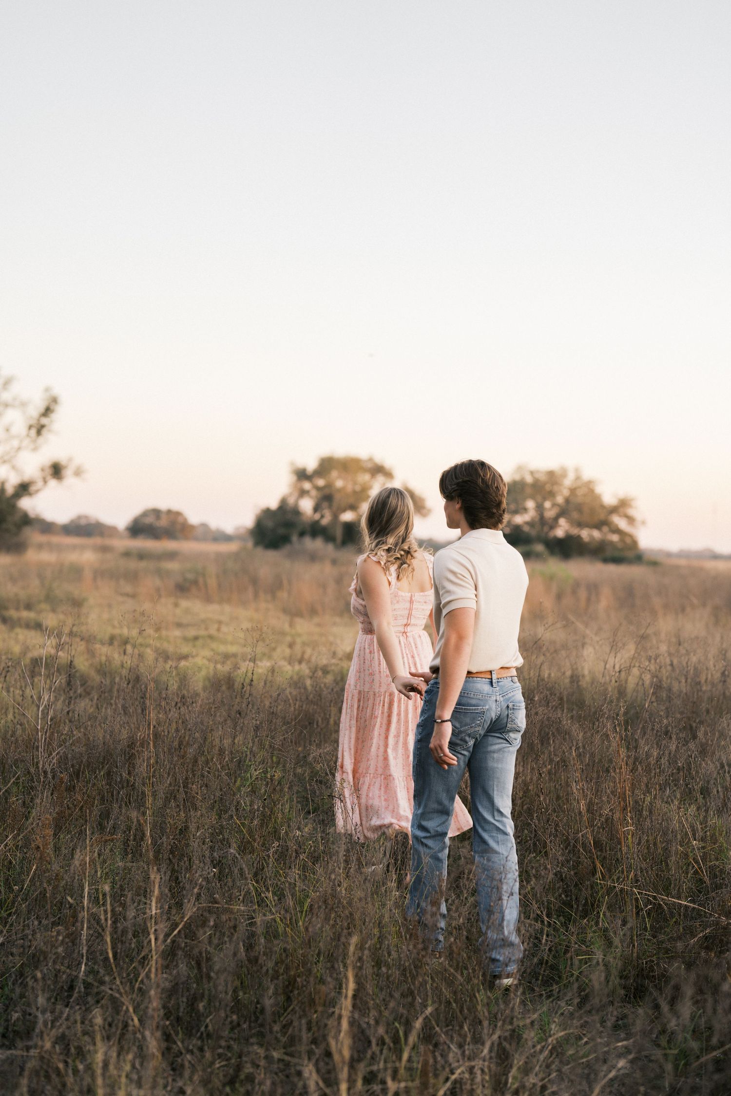 A couple stands together in a golden field at sunset, with her wearing a pink dress and him in jeans and white shirt.