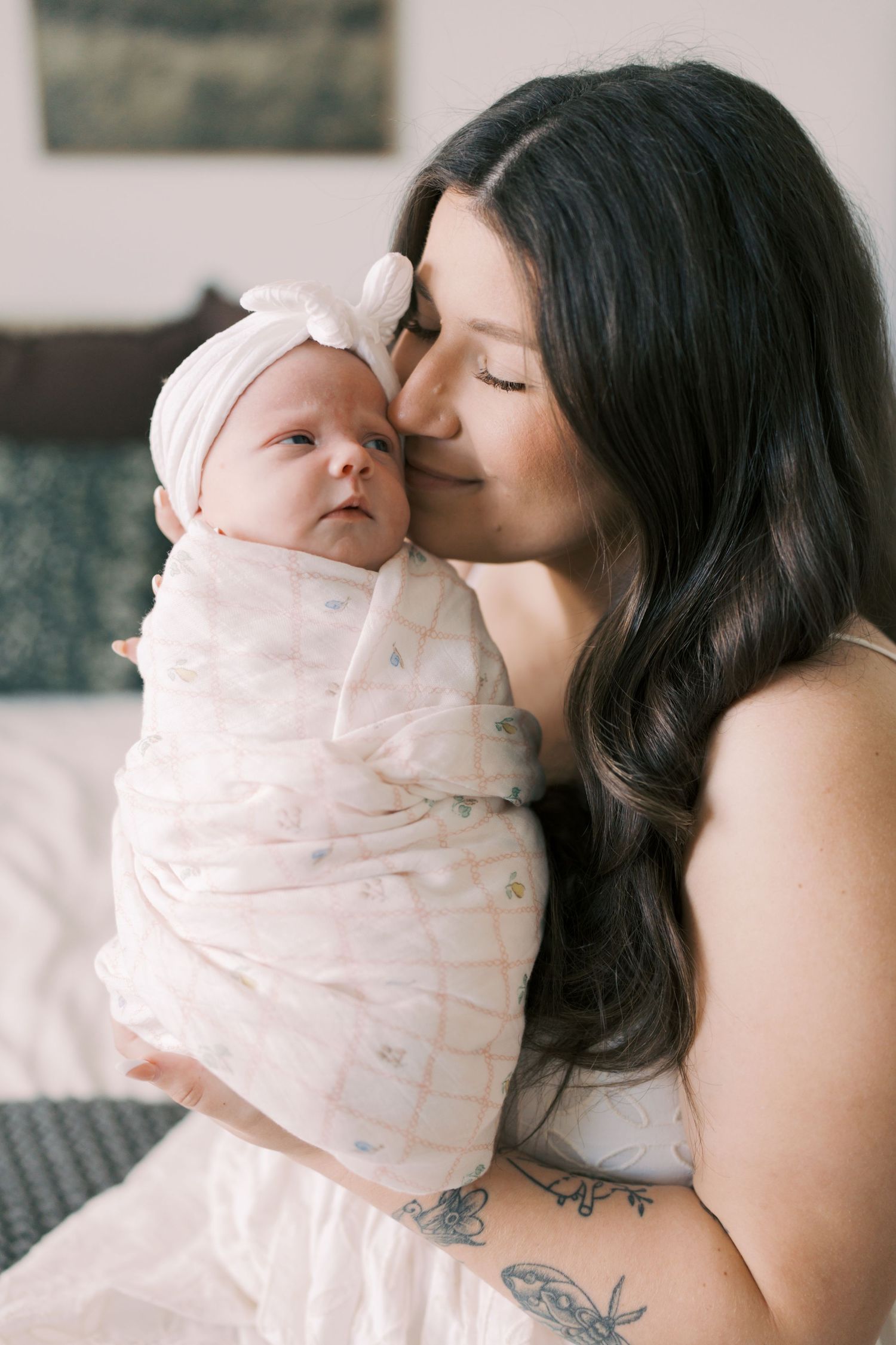 A mother with dark hair gently kisses her swaddled newborn baby girl wearing a white bow headband.