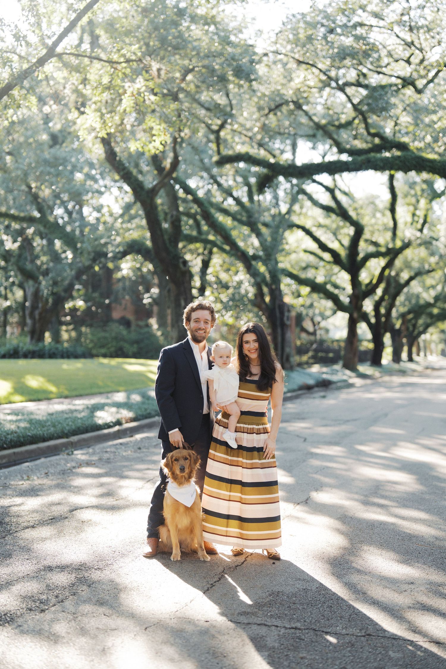 A couple and their dog pose for photos under oak trees on a sunlit path in a park.