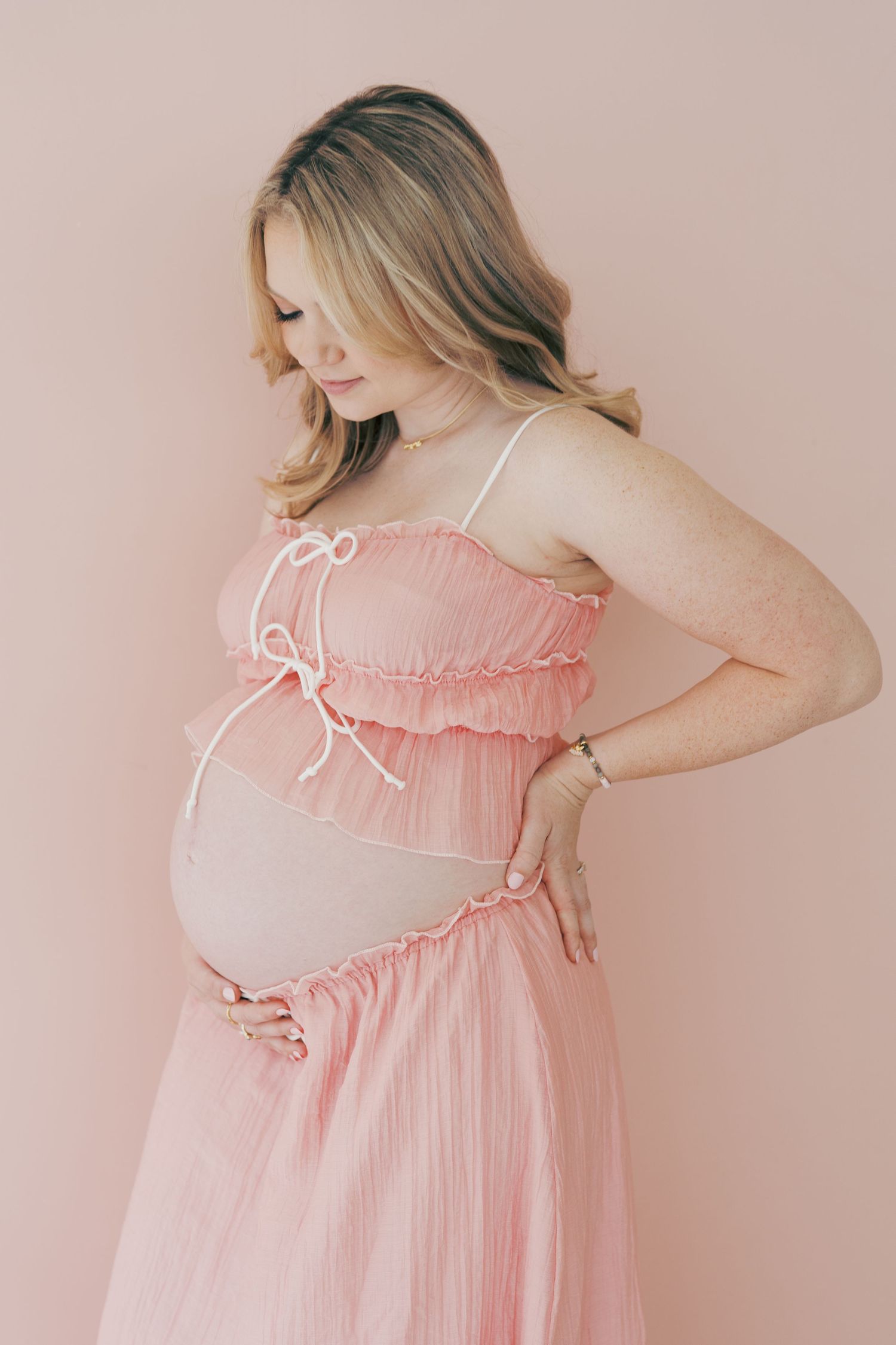 Pregnant woman in pink ruffled maternity dress cradling baby bump against a soft pink background.