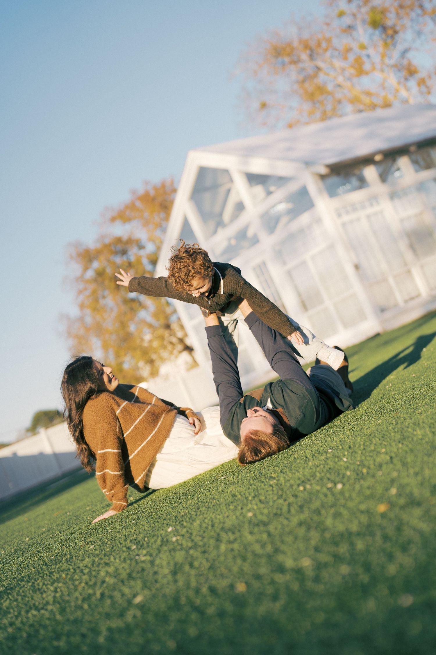 Family playing outdoors on grass, father lifting child in air while mother watches, sunny autumn day.