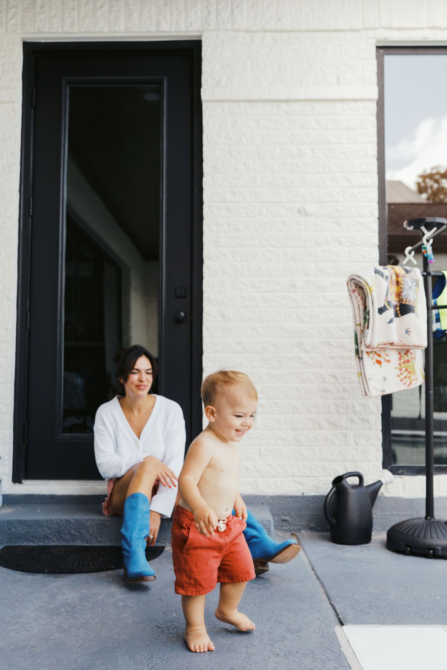 A woman in blue boots sits on front steps watching a shirtless toddler in red shorts playing outside.