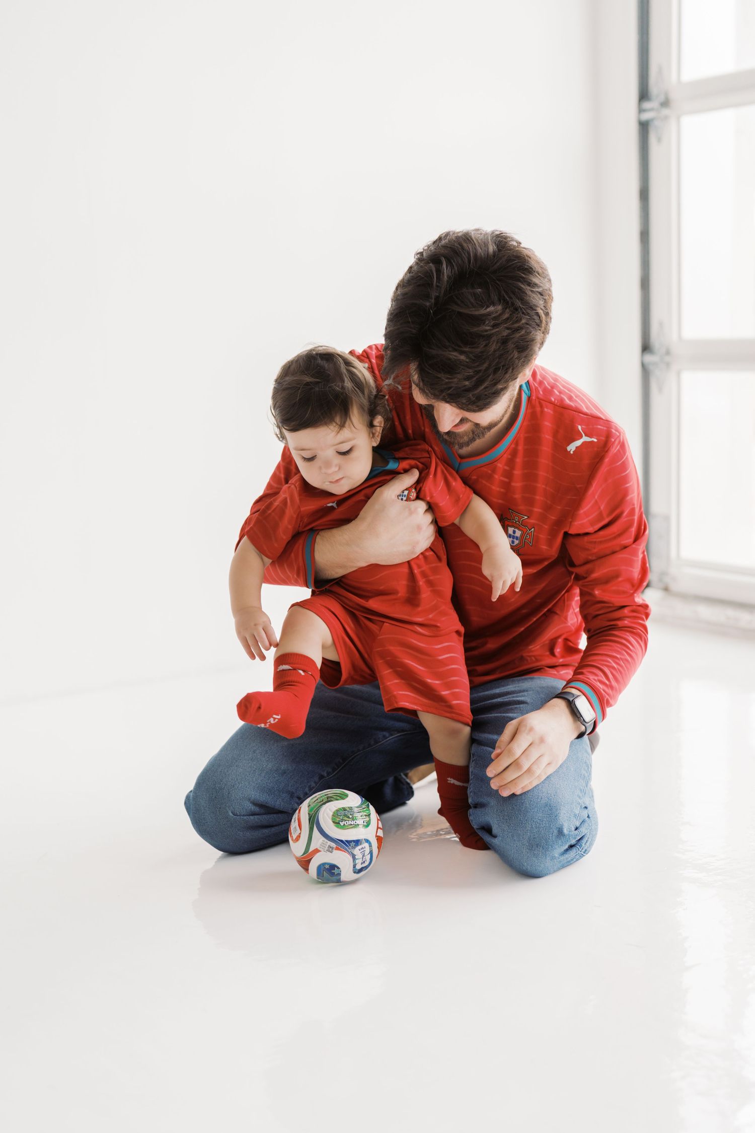 A man is kneeling on the floor and playing with a young child, who is wearing a red outfit.