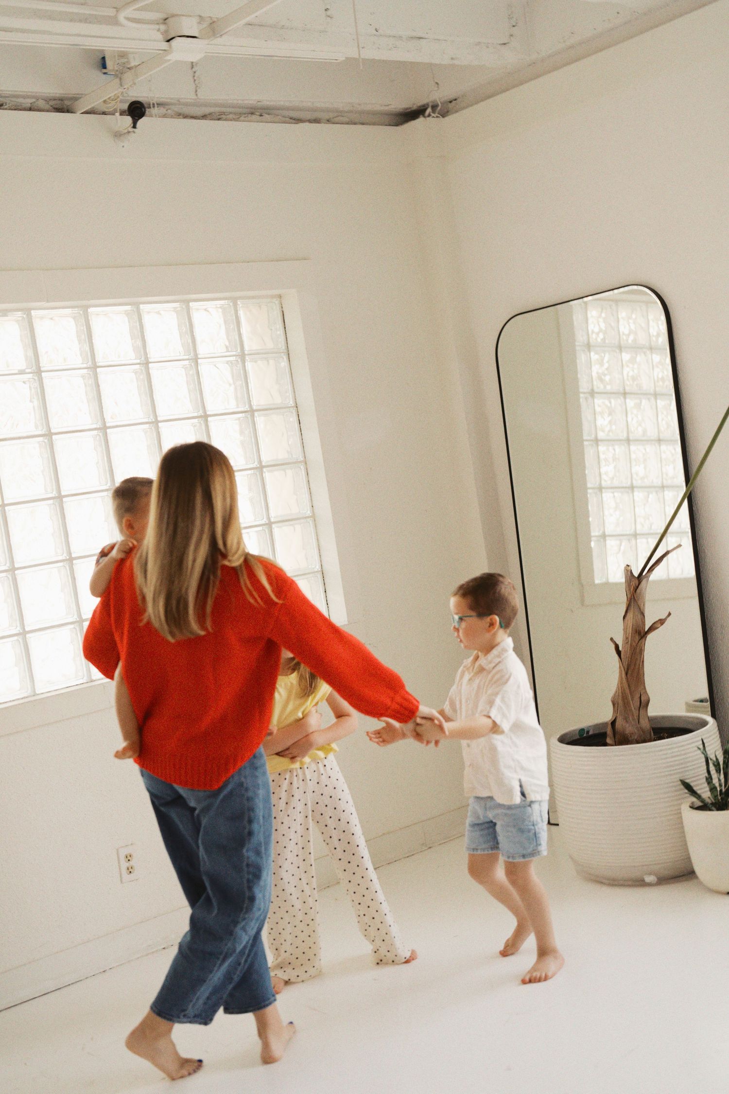A woman in a red sweater dances barefoot with two children in a bright white room with a large mirror.