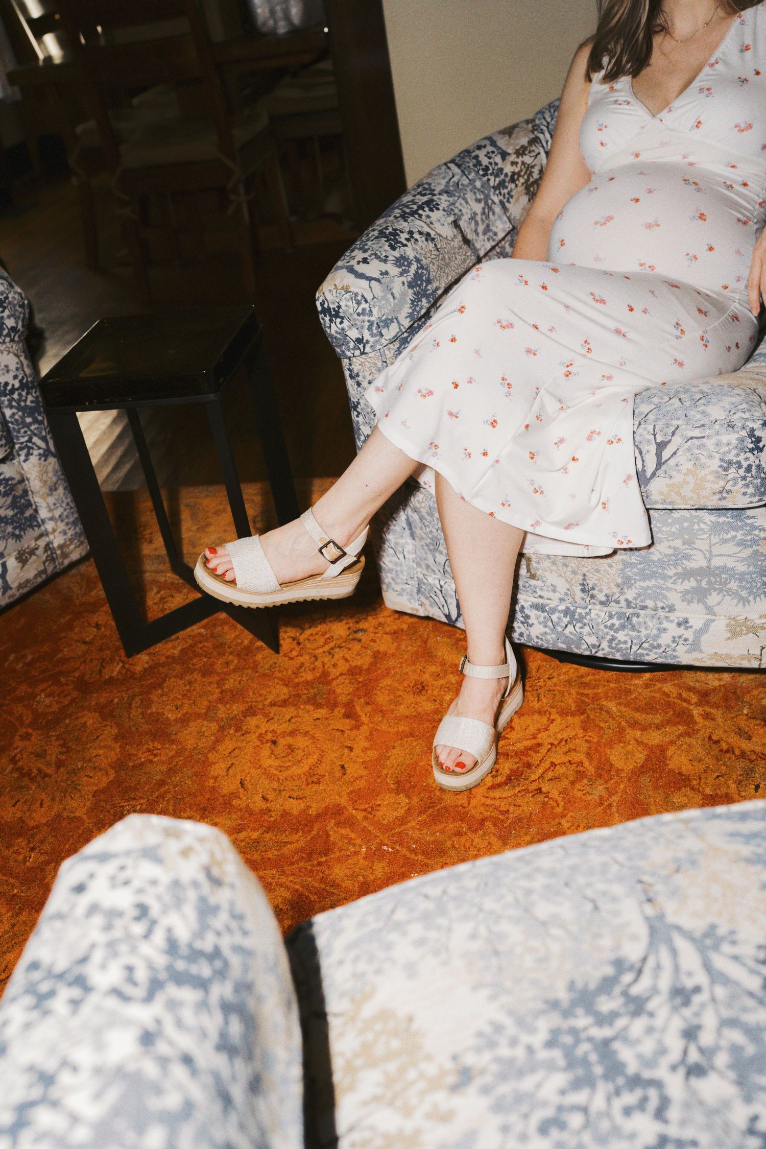 Pregnant woman in floral dress and white platform sandals sitting on blue toile upholstered chair on orange carpet.
