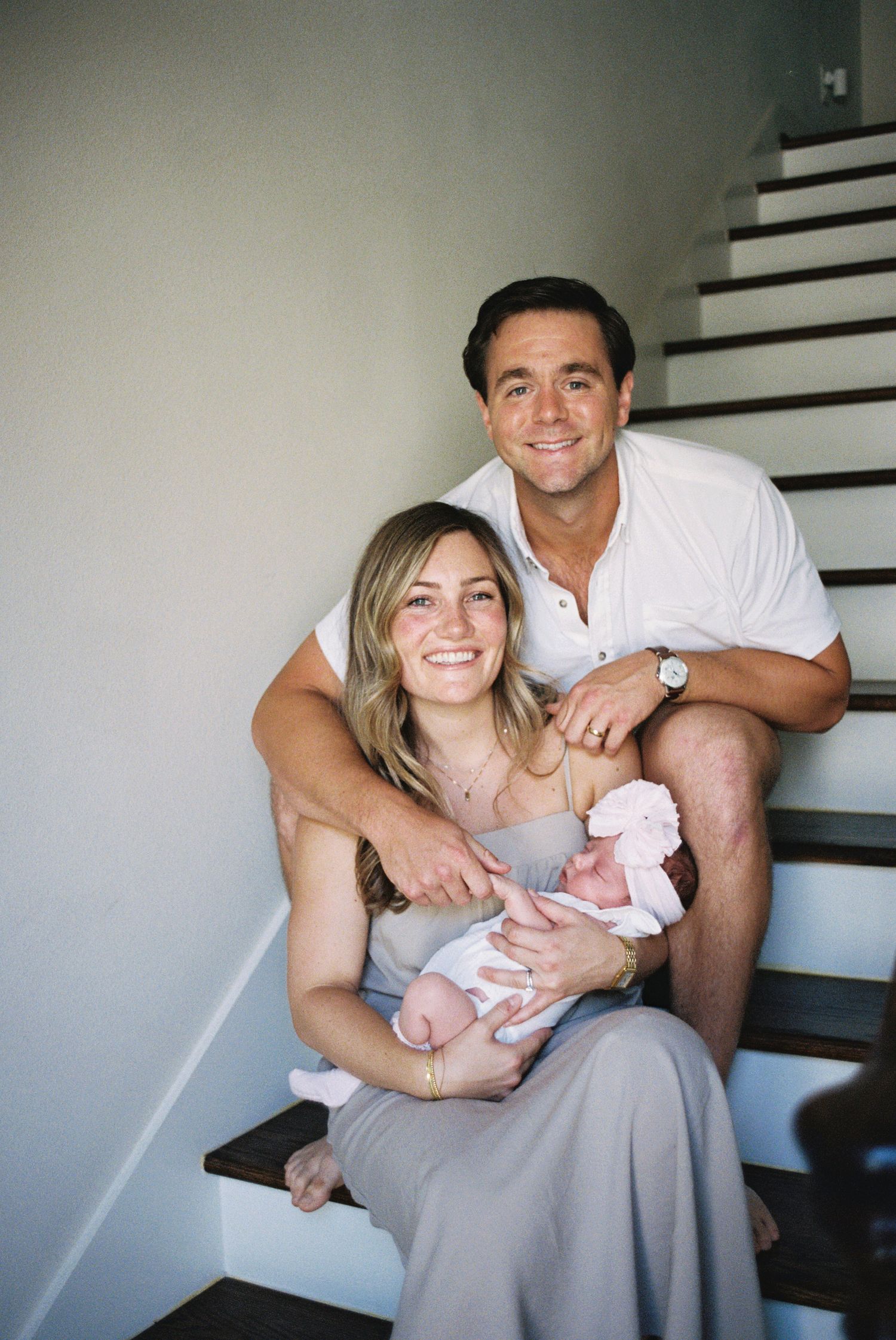 Happy couple sitting on home staircase smiling while holding their newborn baby dressed in pink.