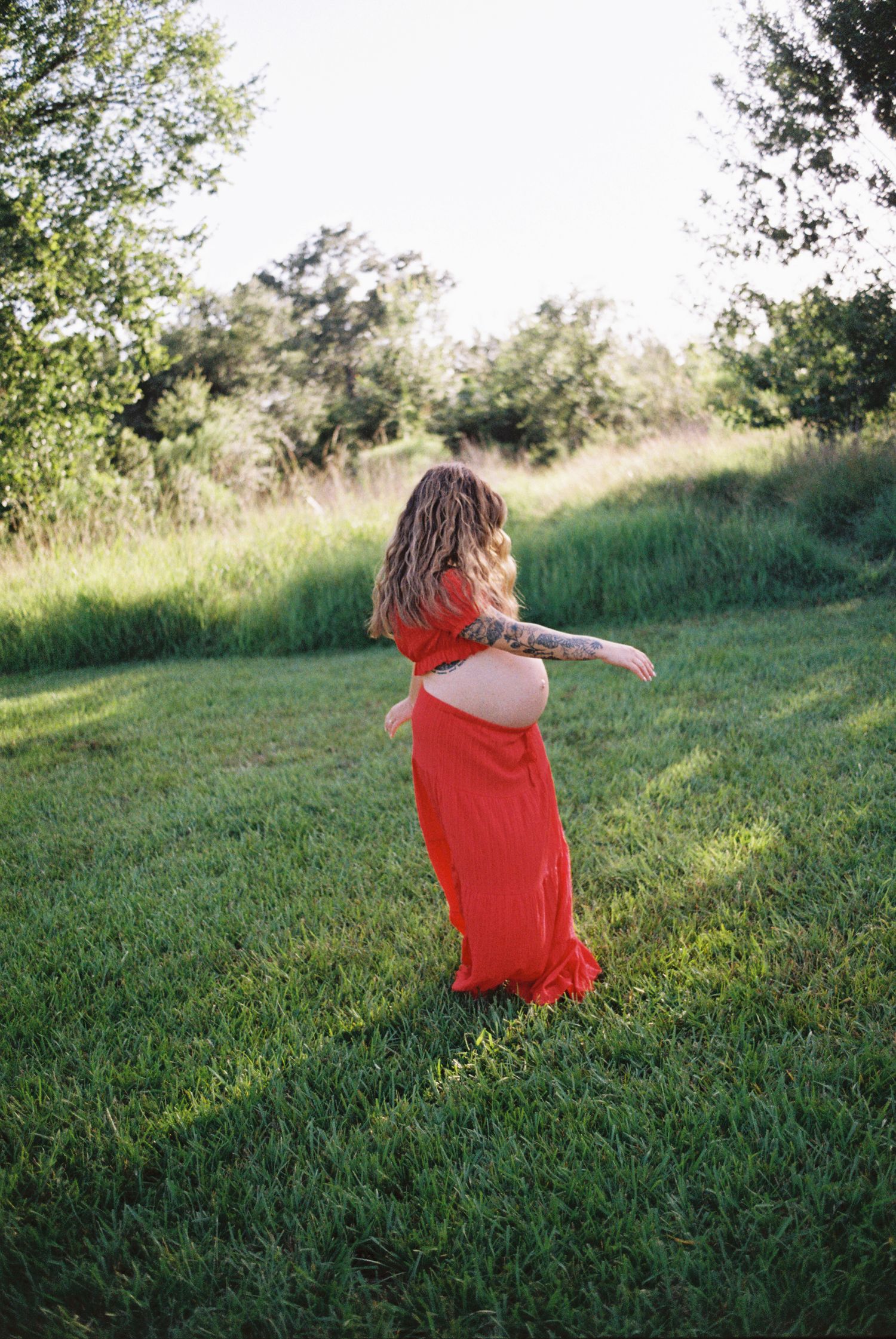 Pregnant woman in red maxi dress with tattoos twirling in lush green field during golden hour maternity photoshoot.