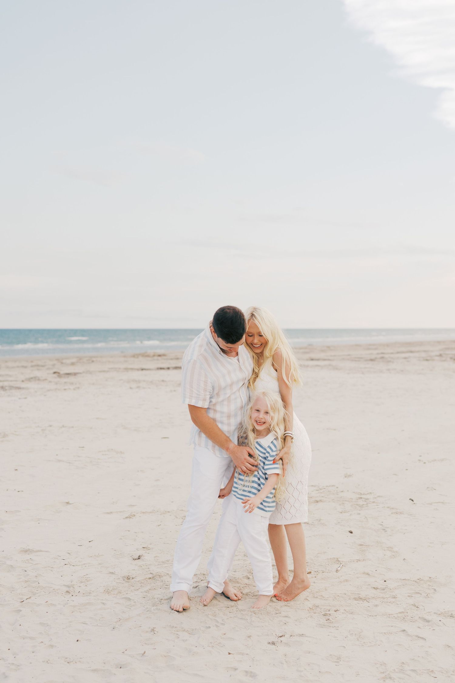 A family enjoys a tender moment together while standing on a sunny beach with ocean waves in the background.