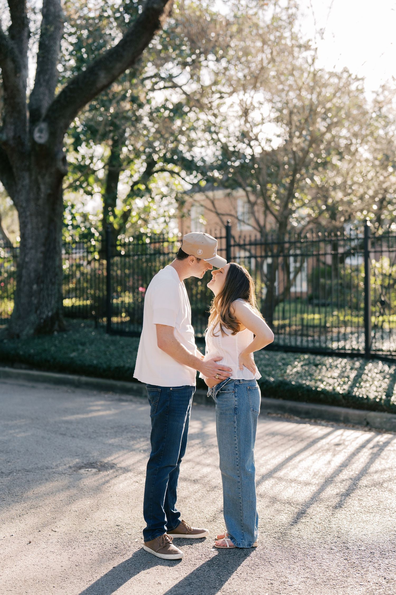 A young couple embracing and smiling in a park setting with trees and fencing in the background.