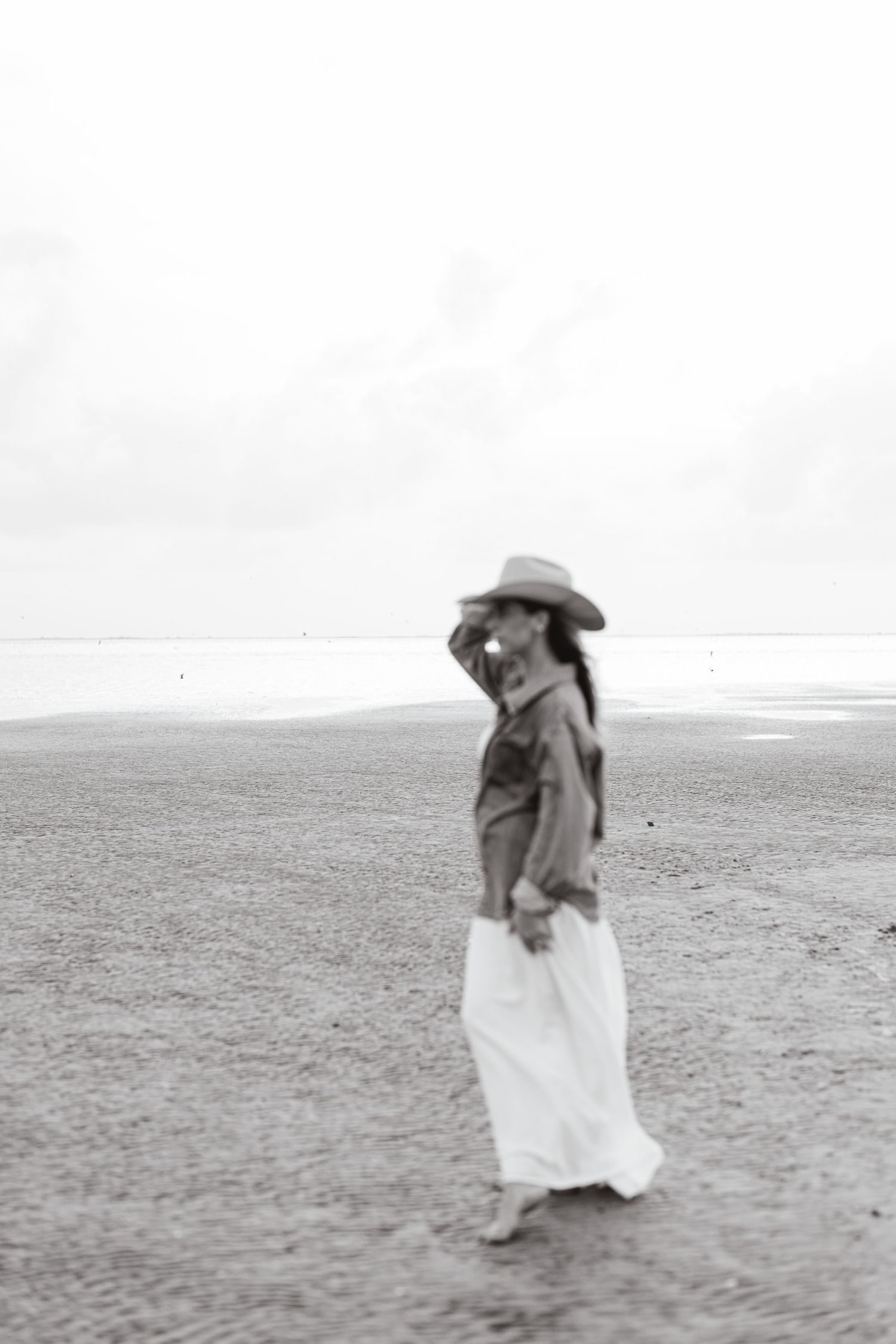 A person in a cowboy hat and long skirt stands alone on a vast empty plain in this vintage black and white photograph.