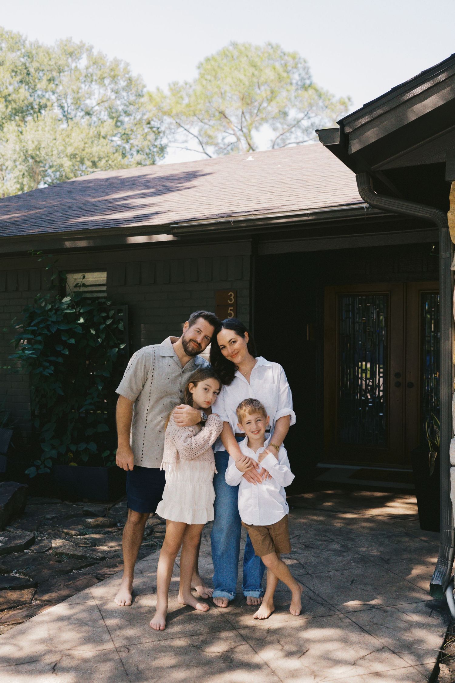 A happy family of four poses barefoot in front of their home, smiling together on a sunny day.