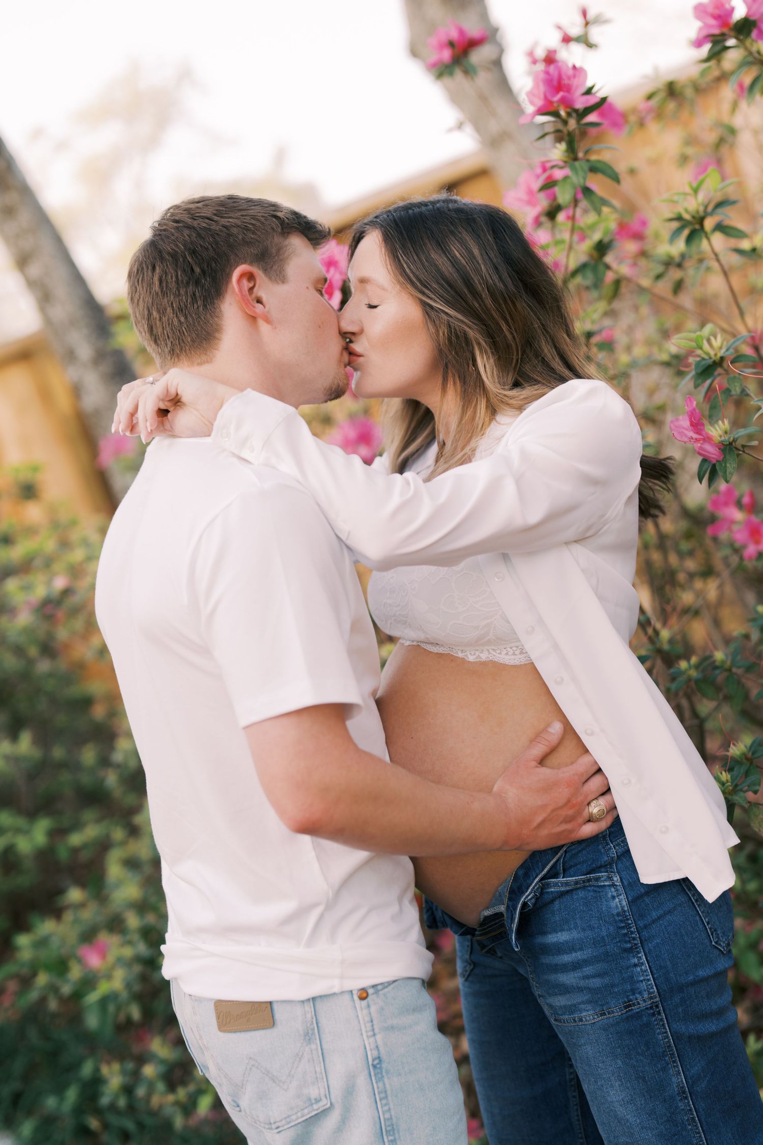 A romantic couple in white tops and jeans share tender kisses against a backdrop of pink flowers in a garden setting.
