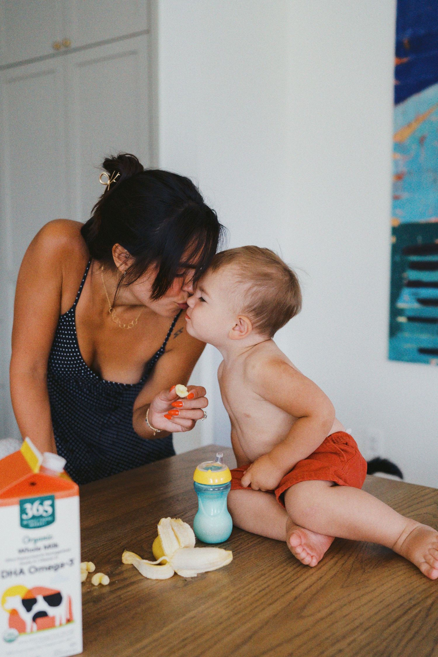 Mother kissing baby sitting on wooden table with banana, sippy cup, and 365 organic whole milk carton nearby.