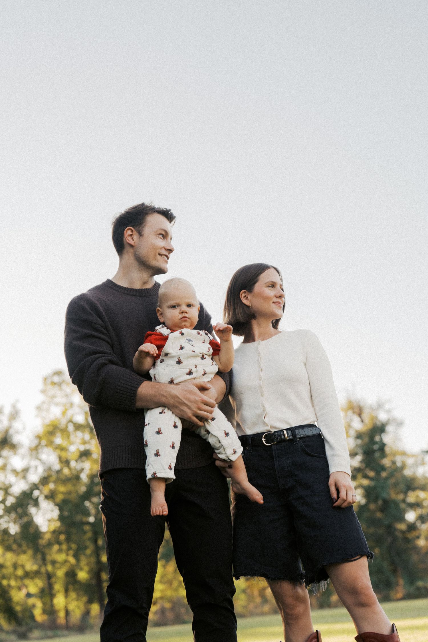 Young family with a baby standing outdoors in a park during golden hour, smiling and enjoying nature.