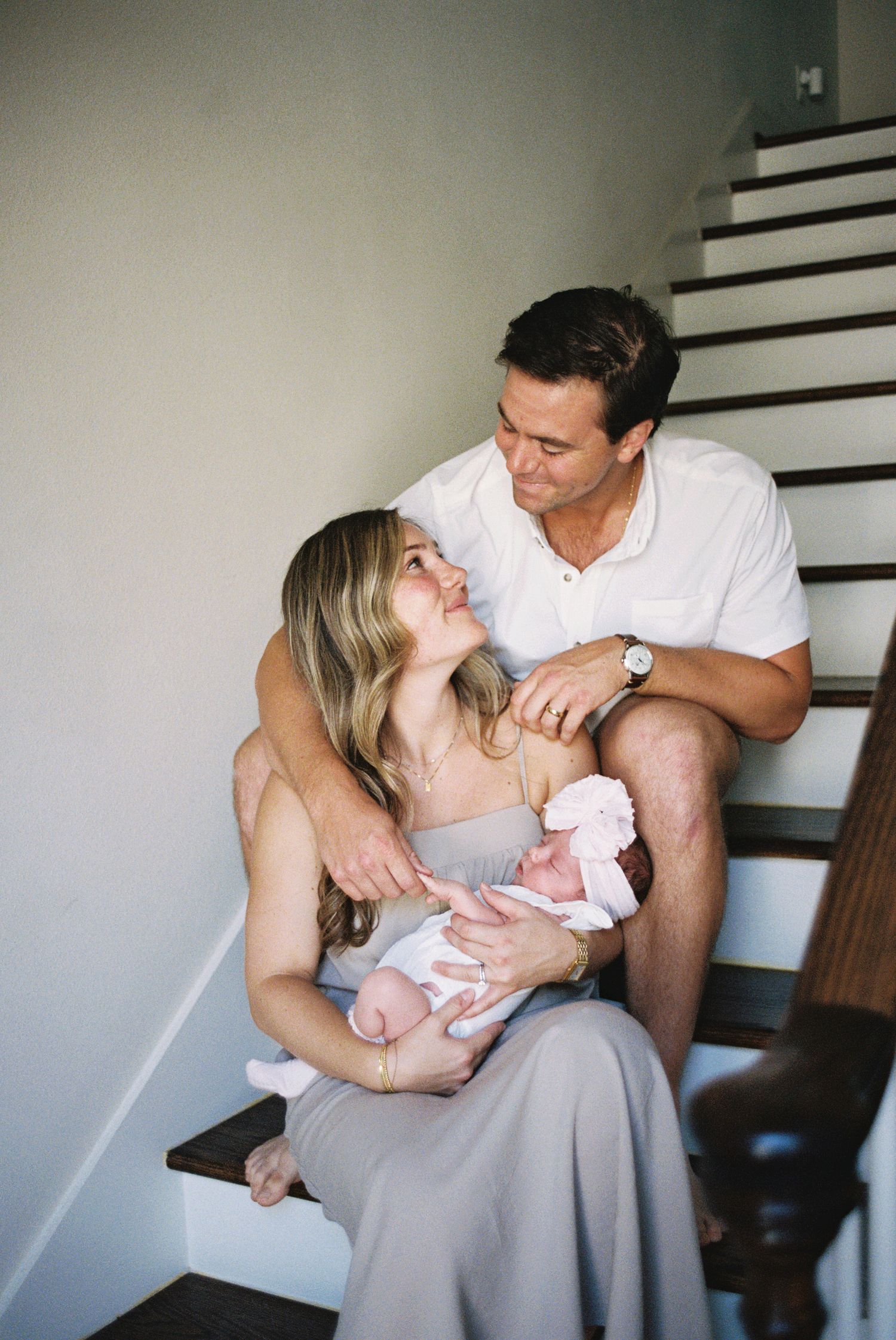 A couple sits on stairs gazing at each other while cradling their newborn baby dressed in pink.