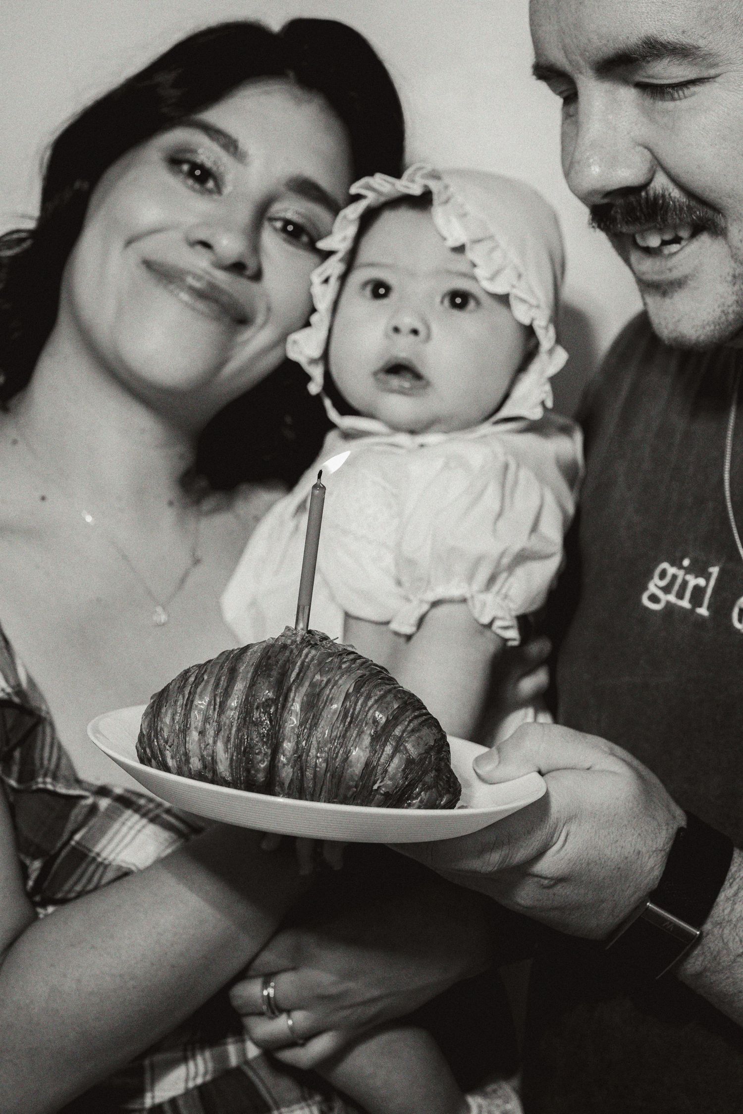 Black and white photo of smiling parents holding baby in bonnet with birthday croissant cake and candle.