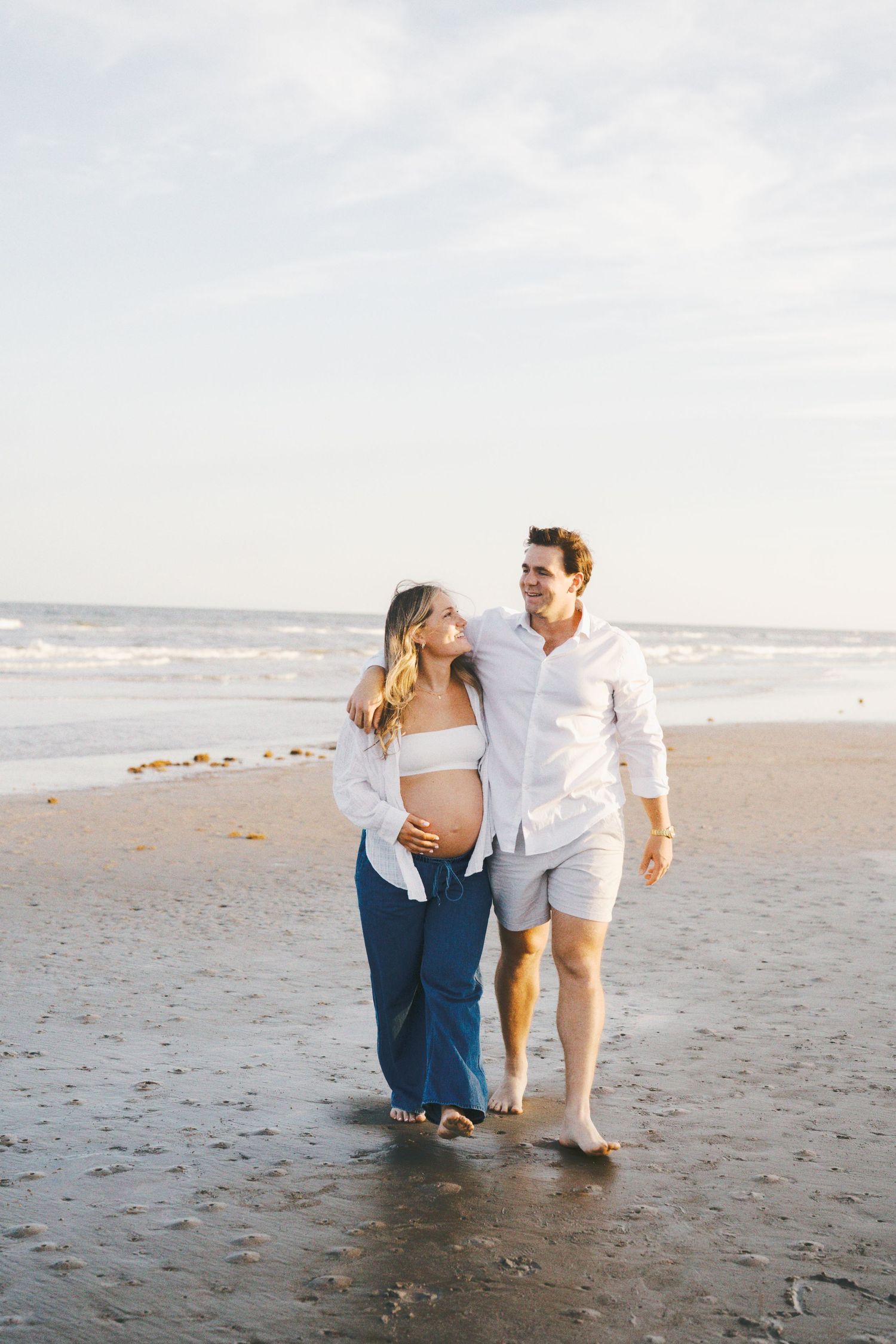 Pregnant couple walking barefoot on beach, woman in blue pants and white top, man in white shirt and shorts.