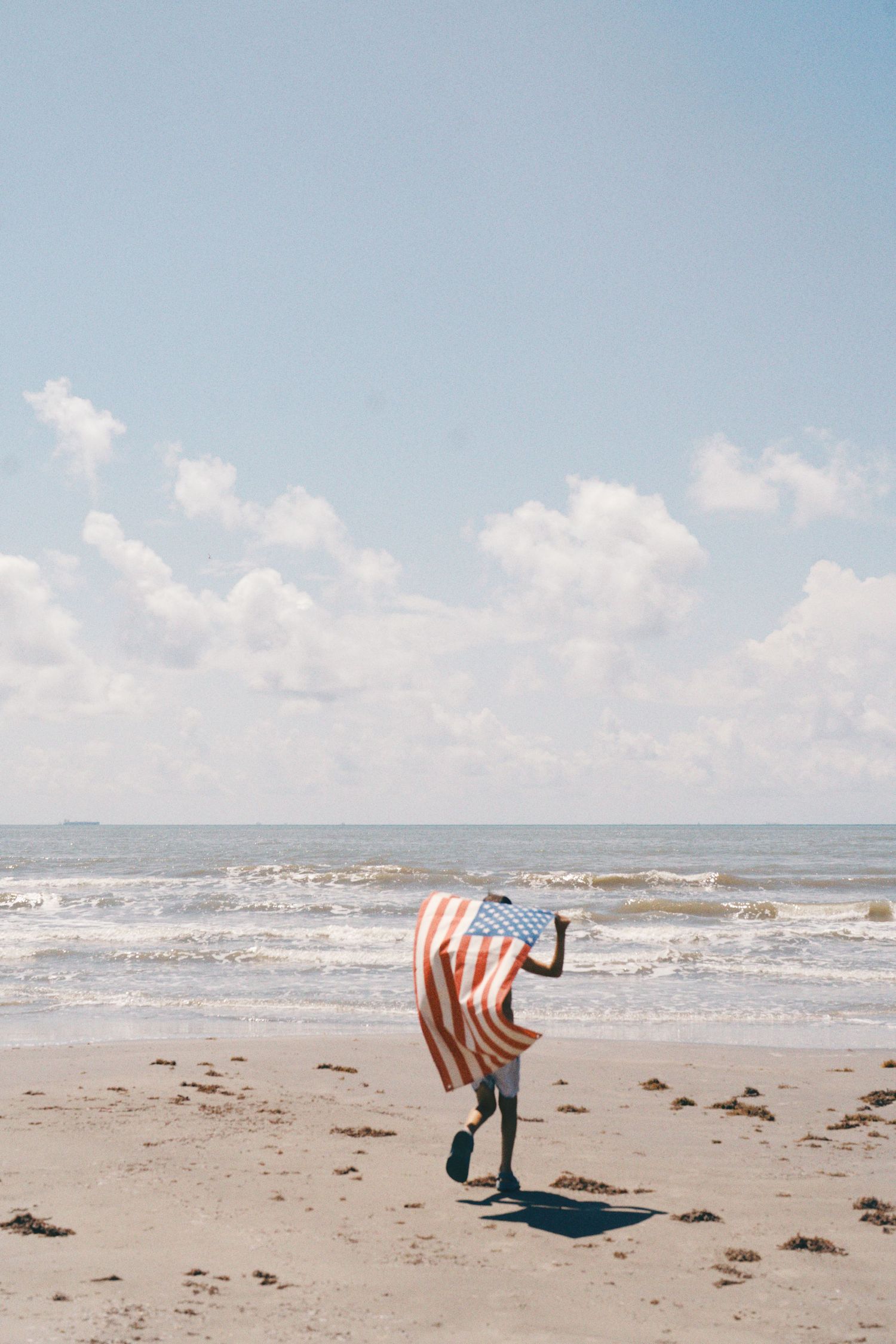 Person running on beach wrapped in American flag with ocean waves and cloudy sky in background.
