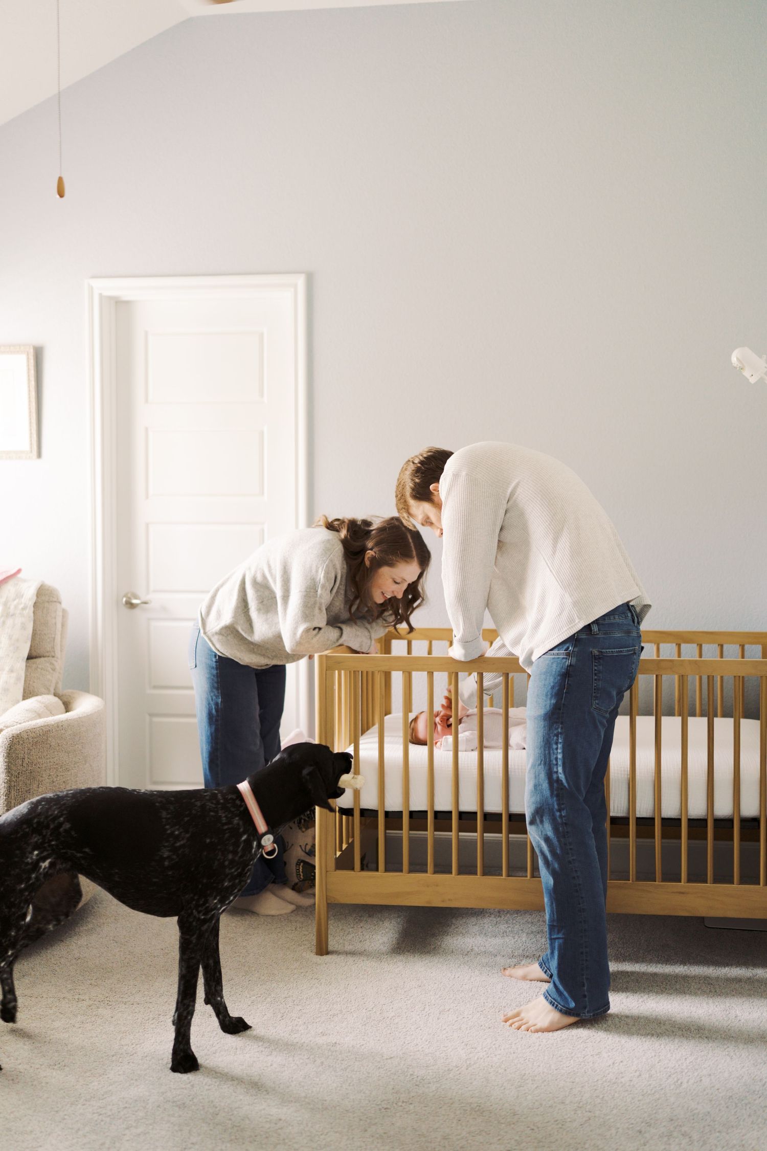 Parents leaning over a wooden crib with their newborn baby, while a black dog watches in a soft nursery.