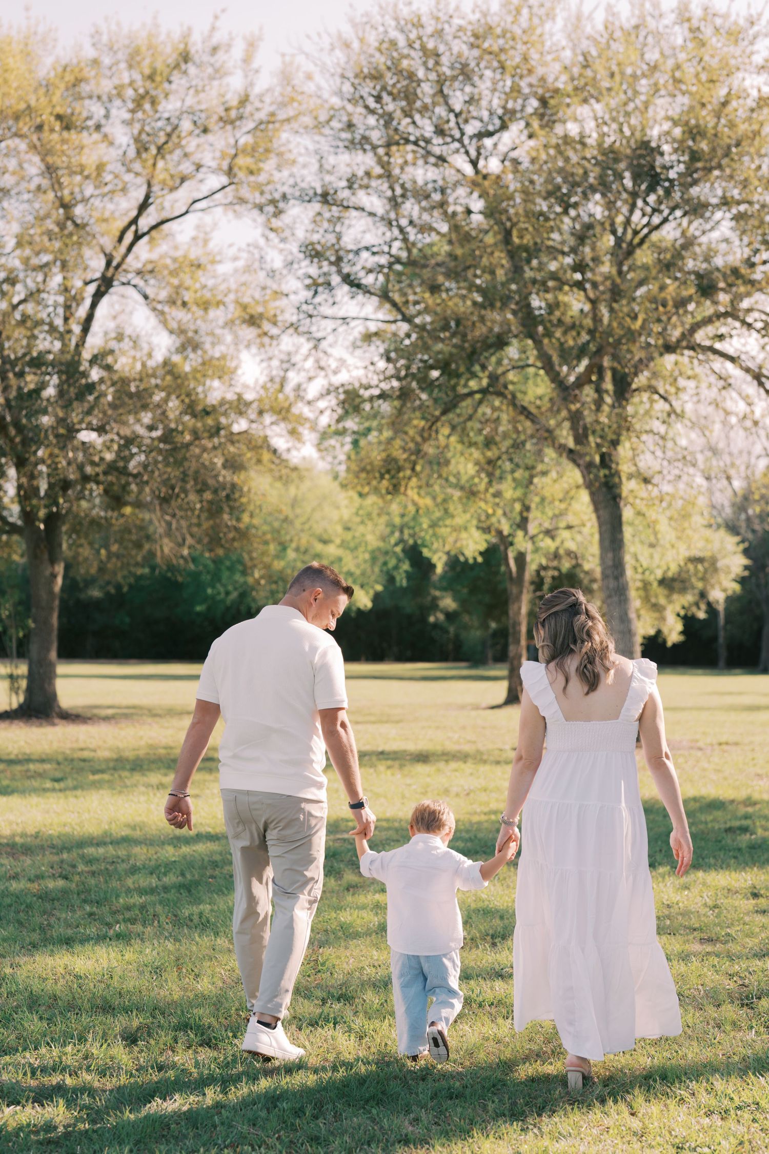 A family walks together through a sun-drenched meadow with tall trees casting gentle shadows in the golden afternoon light.